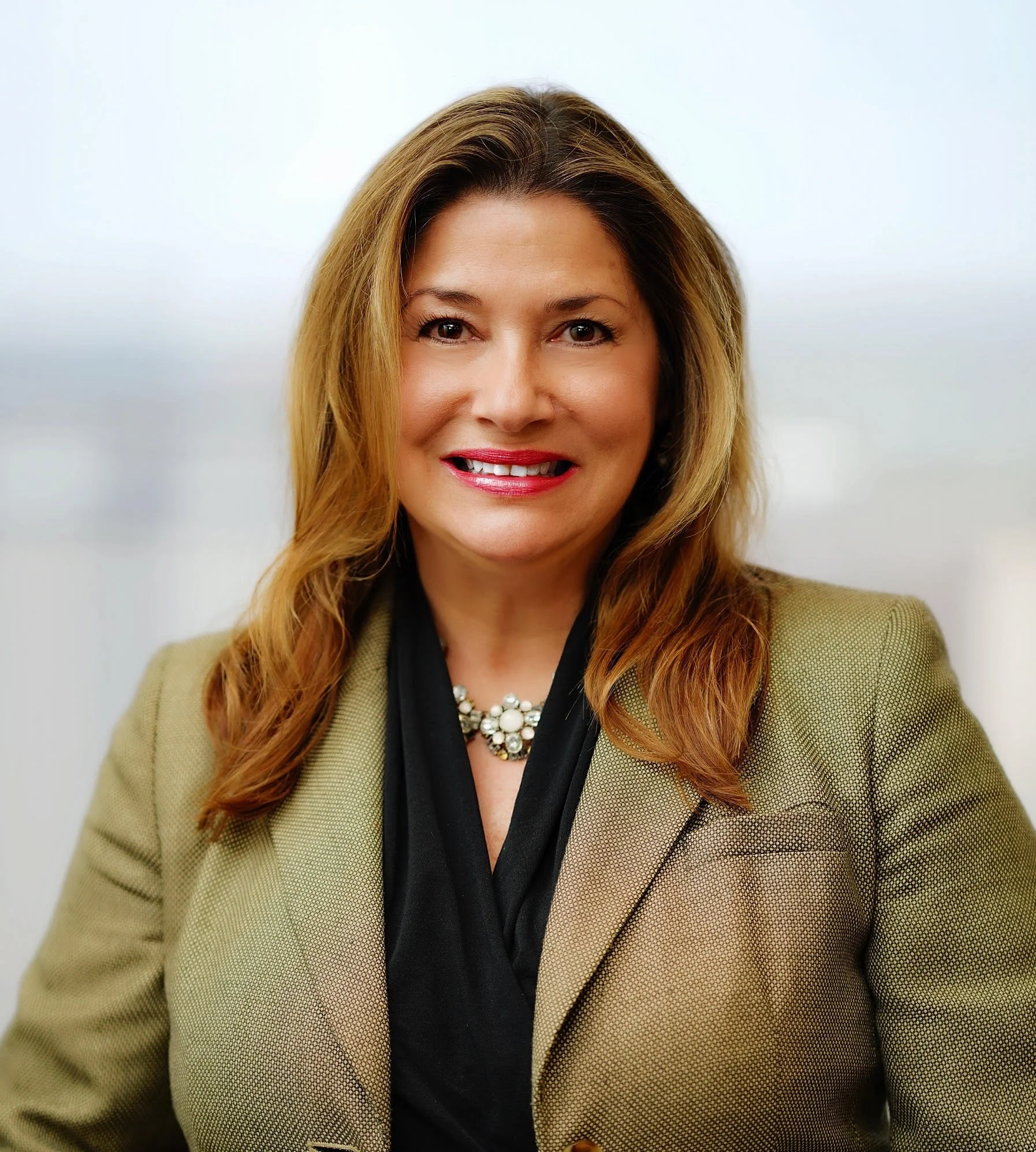 A woman with light skin and shoulder-length reddish-brown hair. She is smiling, wearing a tan blazer, a black top, and a pearl necklace. The background is plain and light-colored.