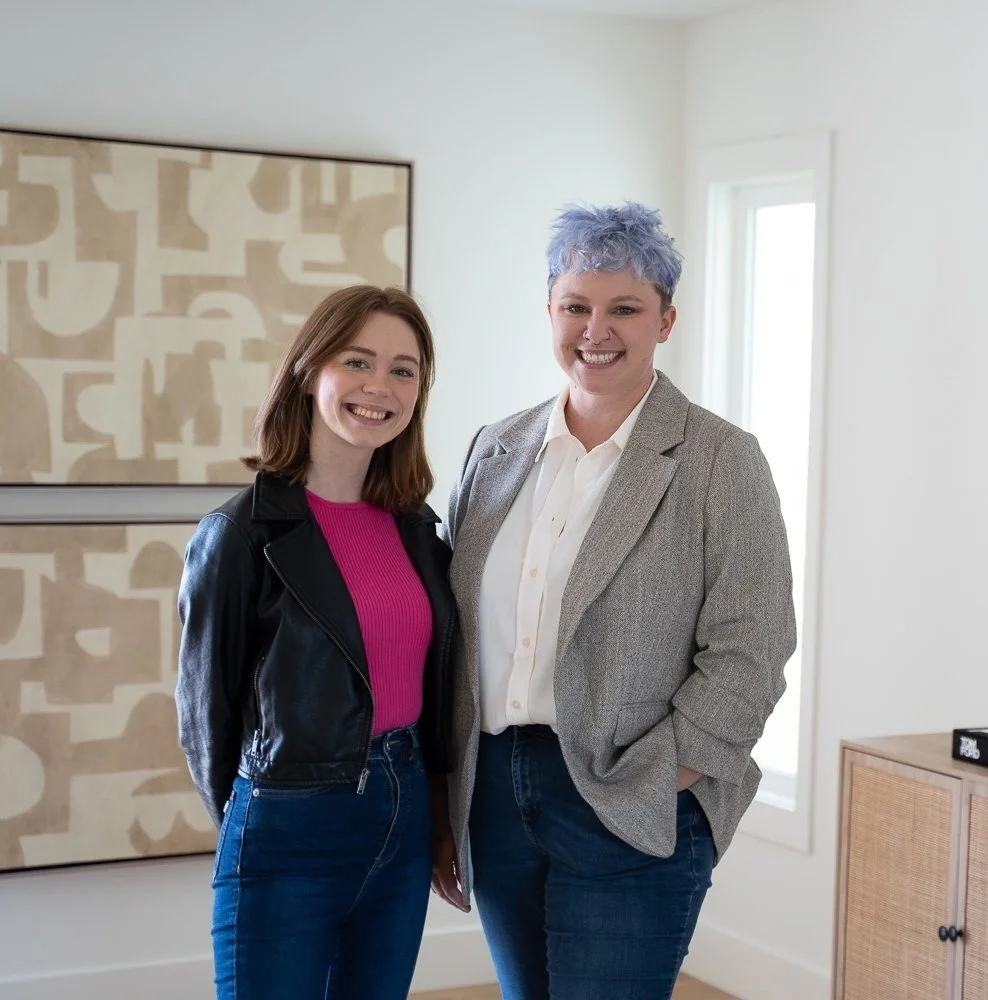 Two women standing indoors, smiling, with one wearing a black leather jacket and pink top, and the other in a gray blazer with a white shirt and blue hair.