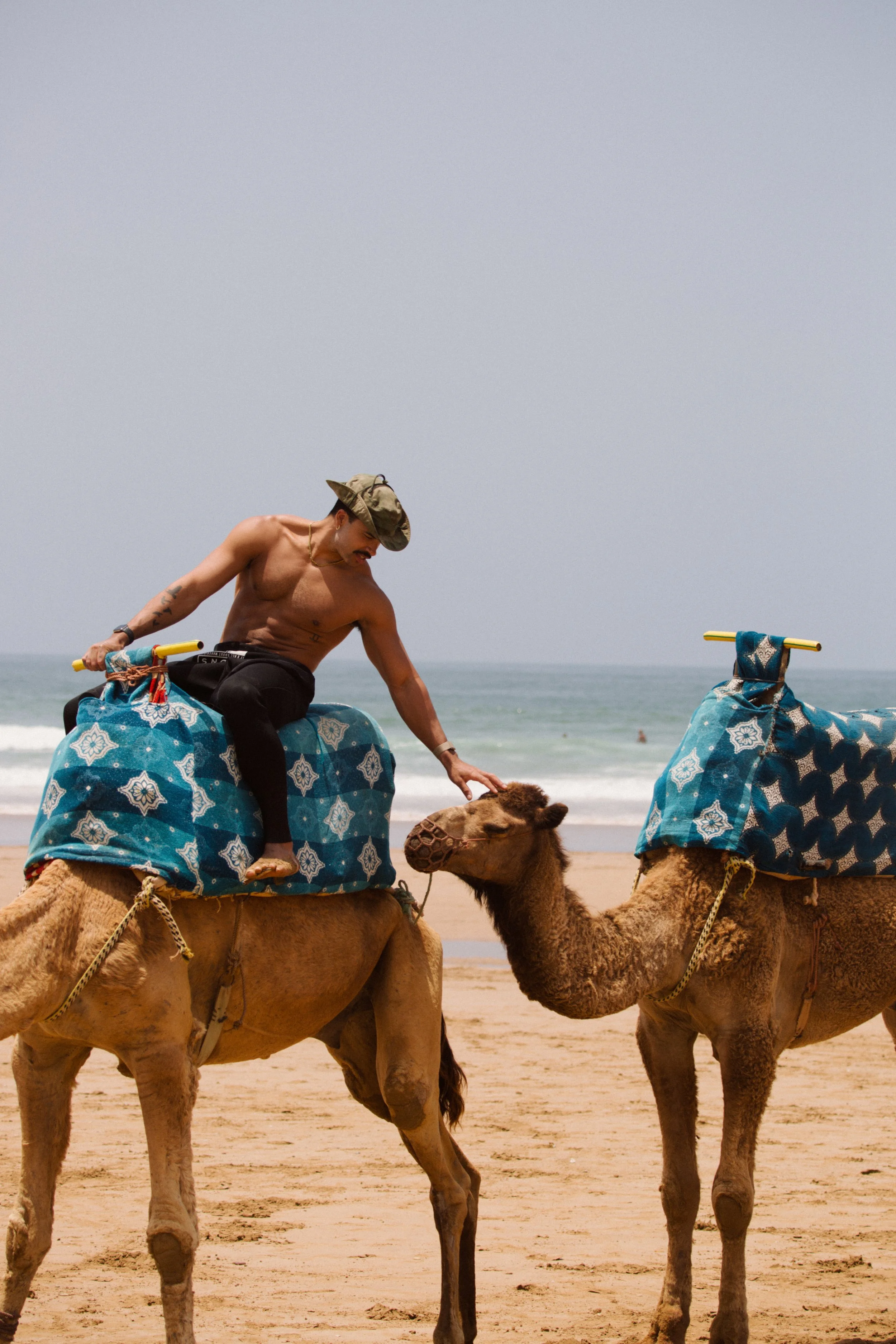 A shirtless man wearing a cap riding a camel on a beach, petting another camel that is standing nearby, with the ocean in the background.