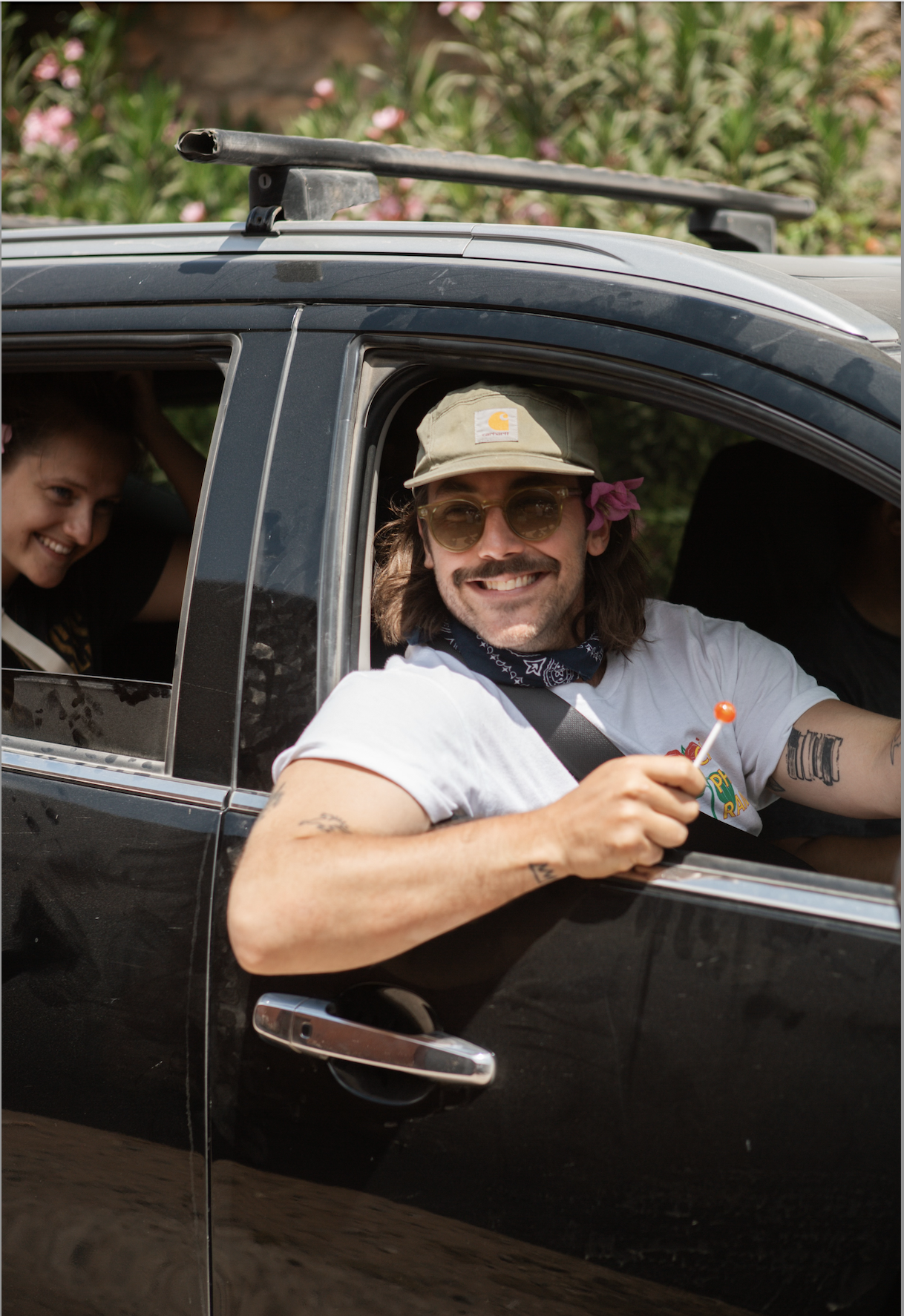 A smiling man with long hair, sunglasses, and a Carhartt hat sitting in the driver’s seat of a black car. He is holding a plastic lollipop and has tattoos on his arms. Another person is partially visible in the backseat, smiling.