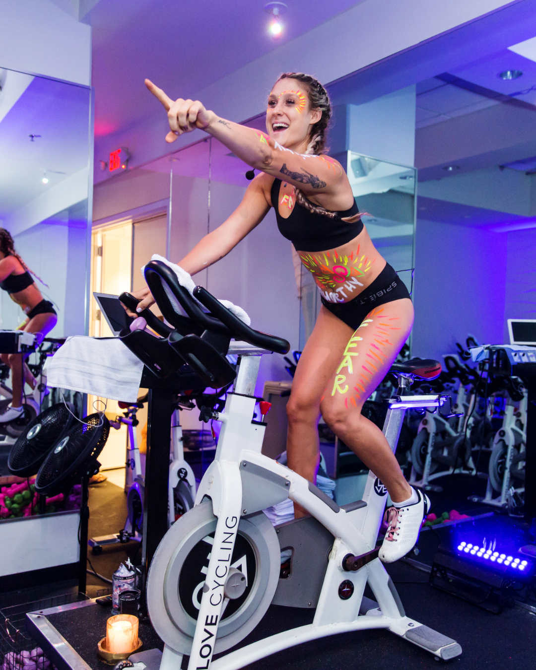 Woman in black workout clothes with colorful body paint, animated expression, and braids, riding a stationary bike in a fitness studio with purple lighting.