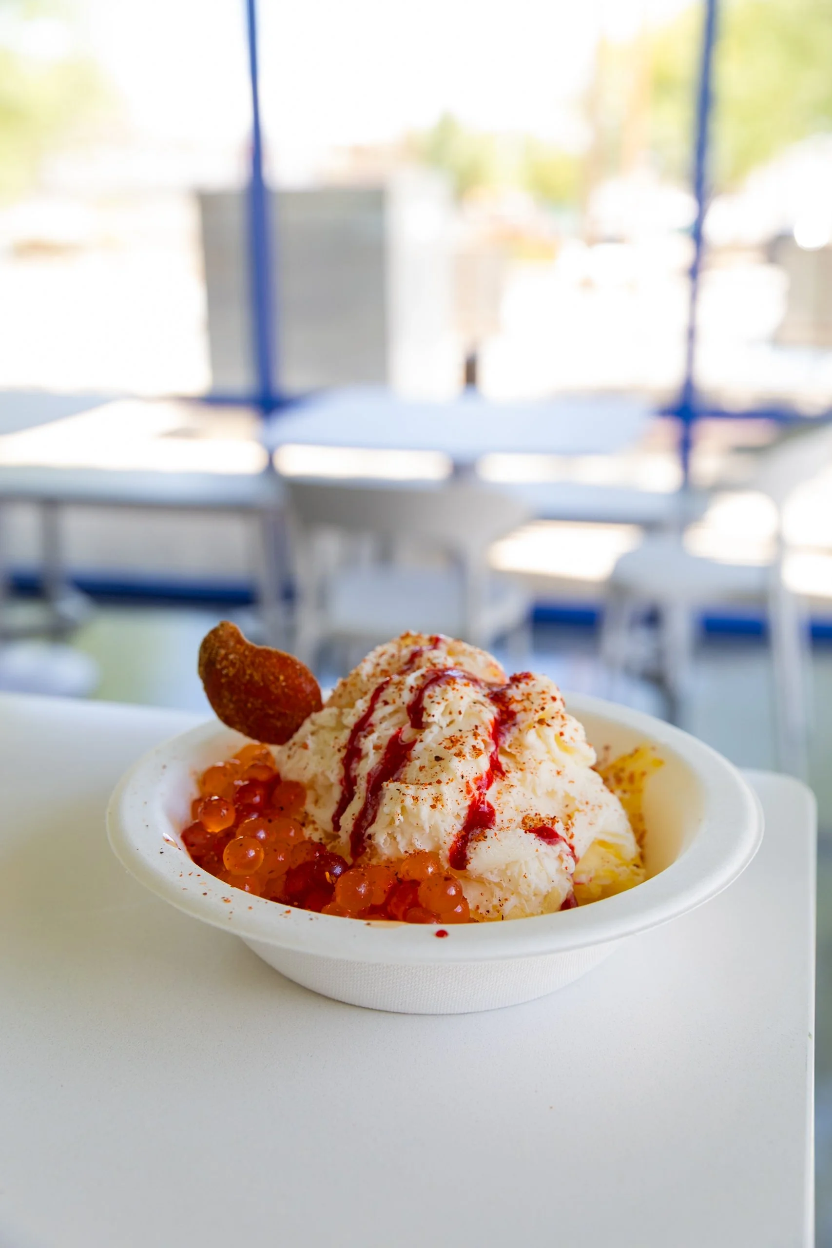 A bowl of mashed ice cream topped with red syrup, surrounded by orange fish roe, with a fried garnish on the side, on a white table in a bright setting.