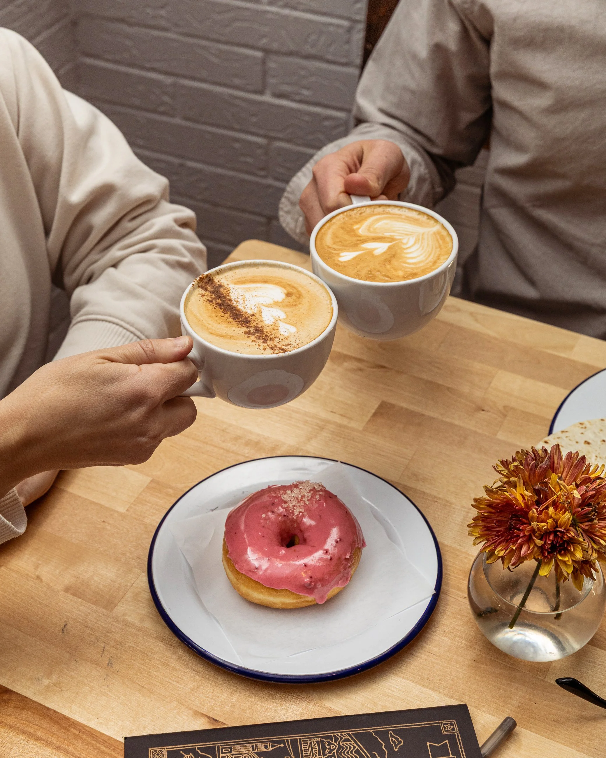 Two people holding cups of coffee with latte art, a pink frosted donut on a plate, and a small flower vase on a wooden table.
