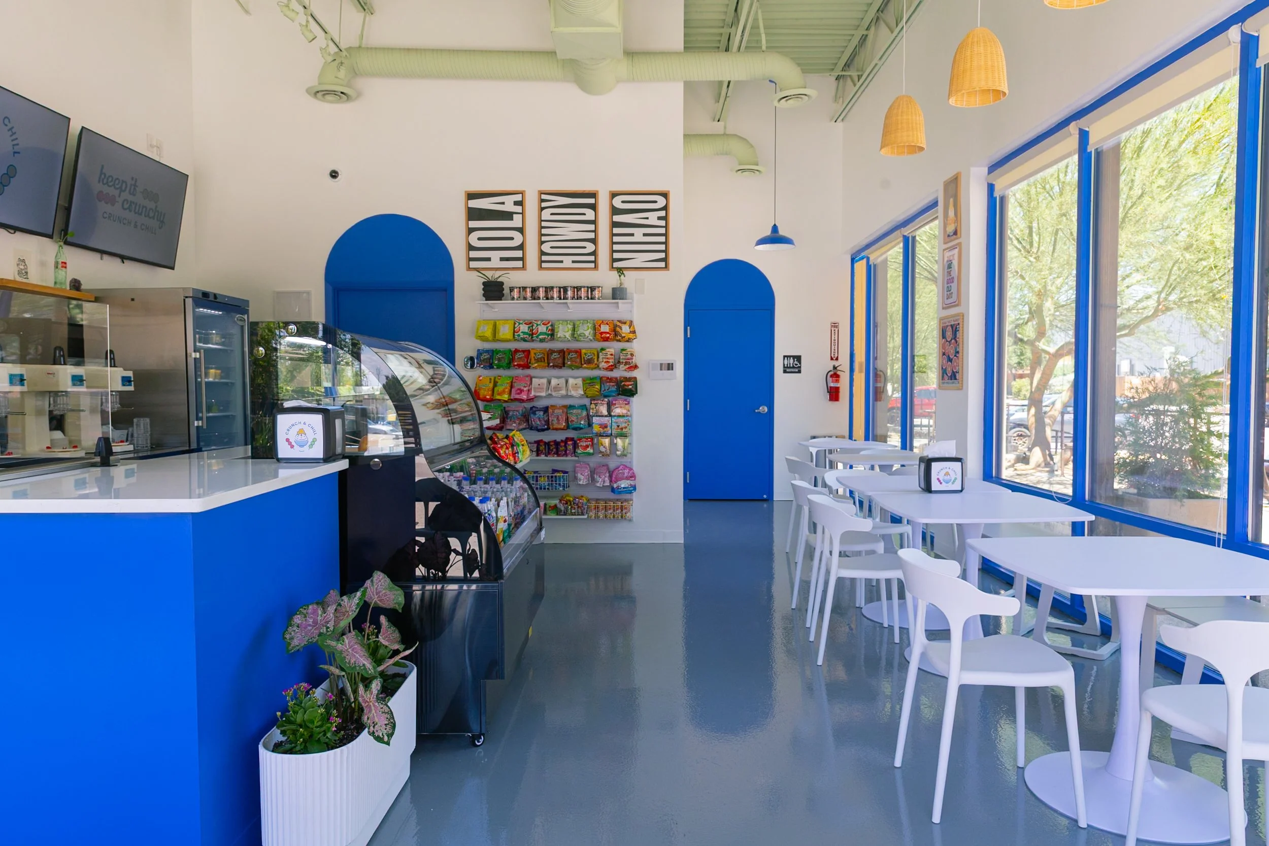 Interior of a bright, modern cafe with white and blue decor, featuring a counter with refrigerated display case, snack shelves, white tables with chairs, large windows, and hanging light fixtures.