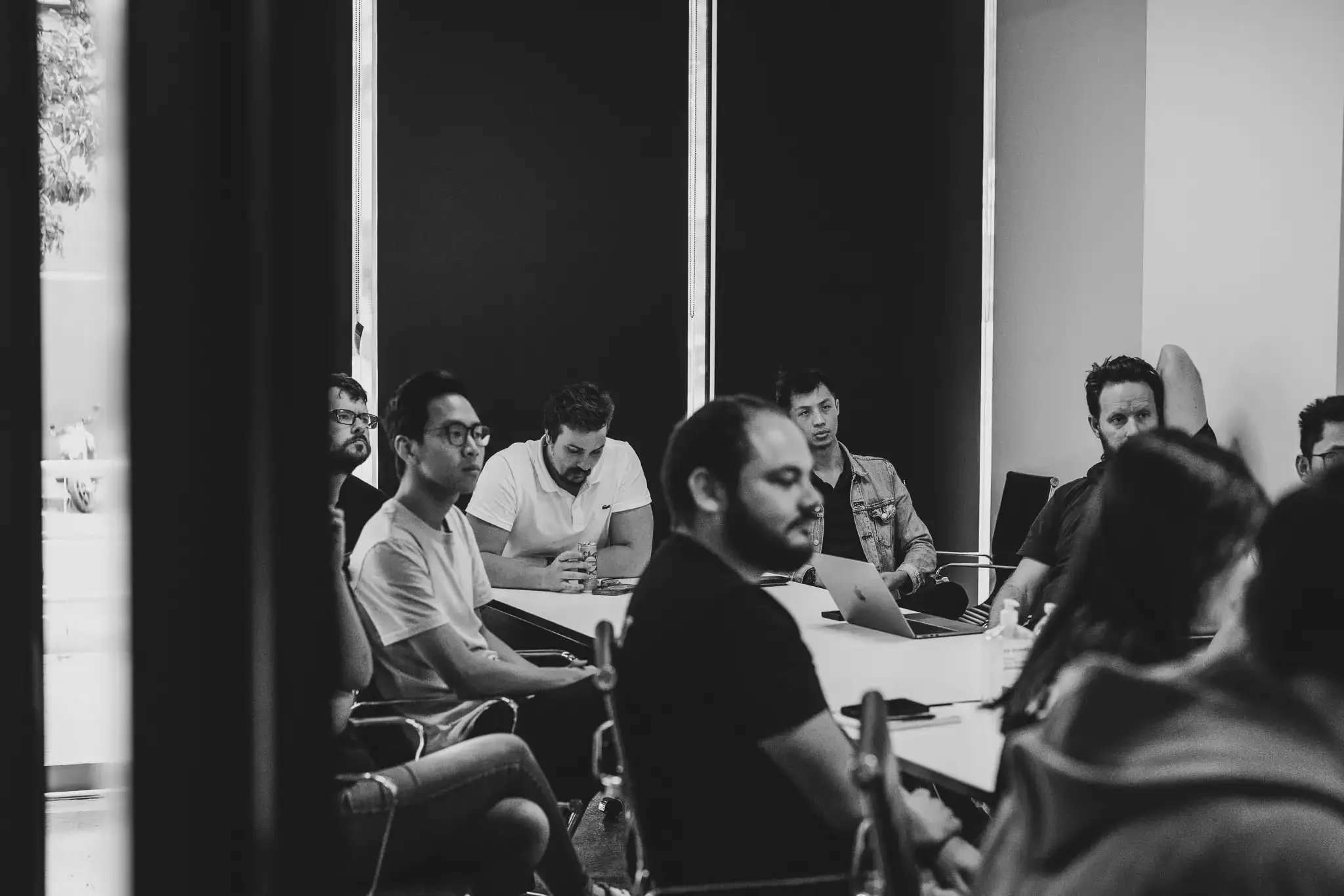 The team (10 people) sitting down in a meeting, all engaged in a conversation or looking at a screen that isn't visible from the photo. They are seated at a white table.