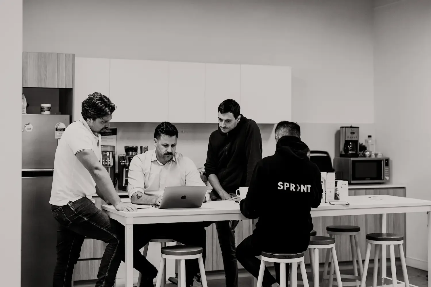 Four men gathered around a kitchen counter, two are sitting, one is standing, and one is leaning, looking at a laptop, in a modern kitchen with white cabinets and appliances.