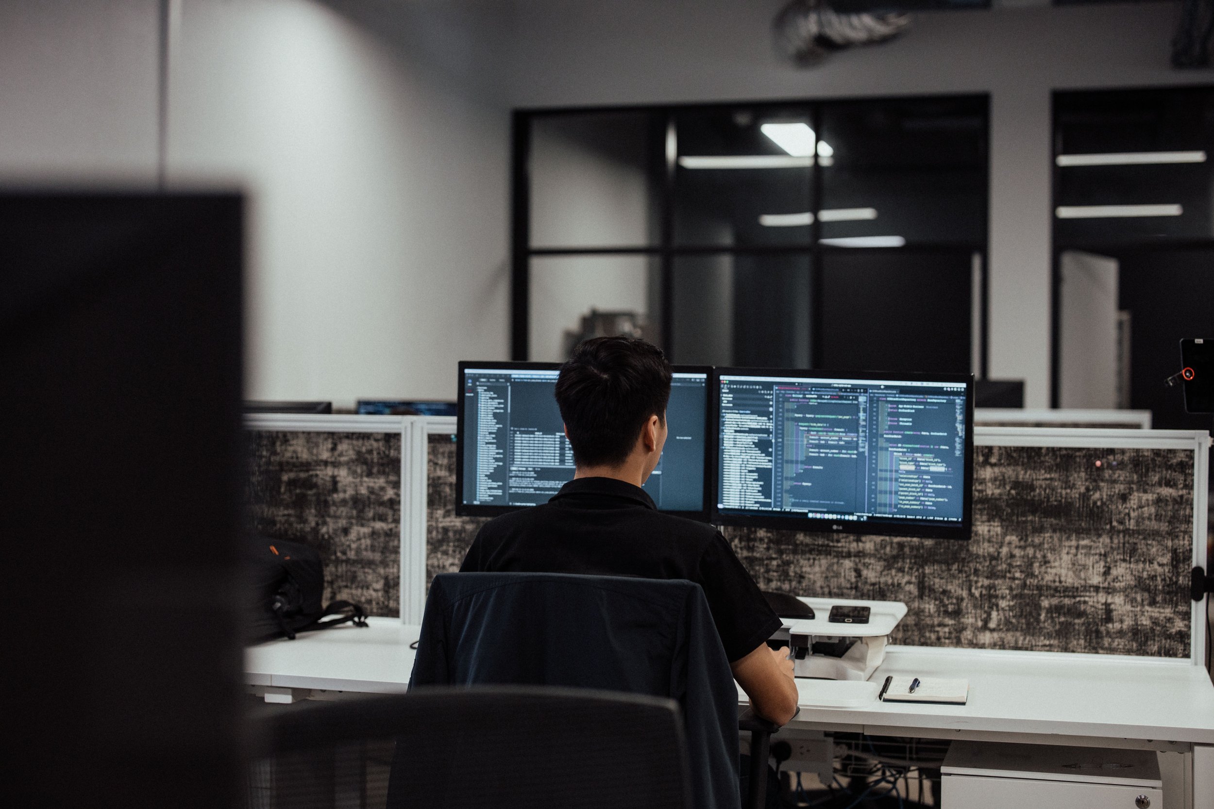 Person working at a dual-monitor computer setup in an office environment, coding visible on screens.