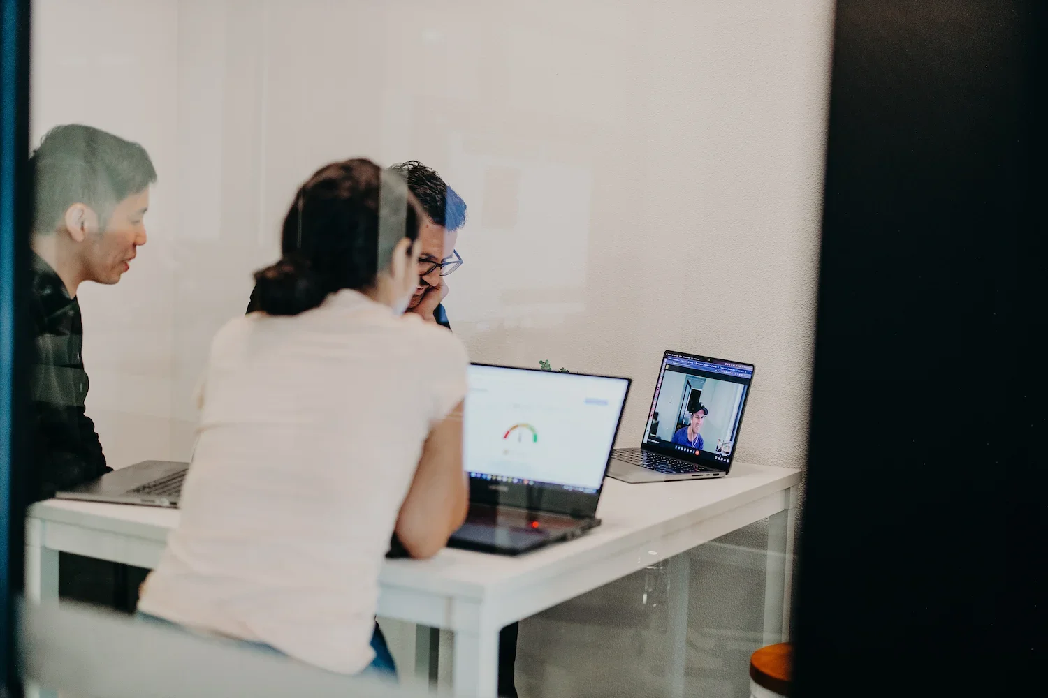 3 people in a meeting room, the photo taken through the glass and they are in a meeting with someone on the laptop screen