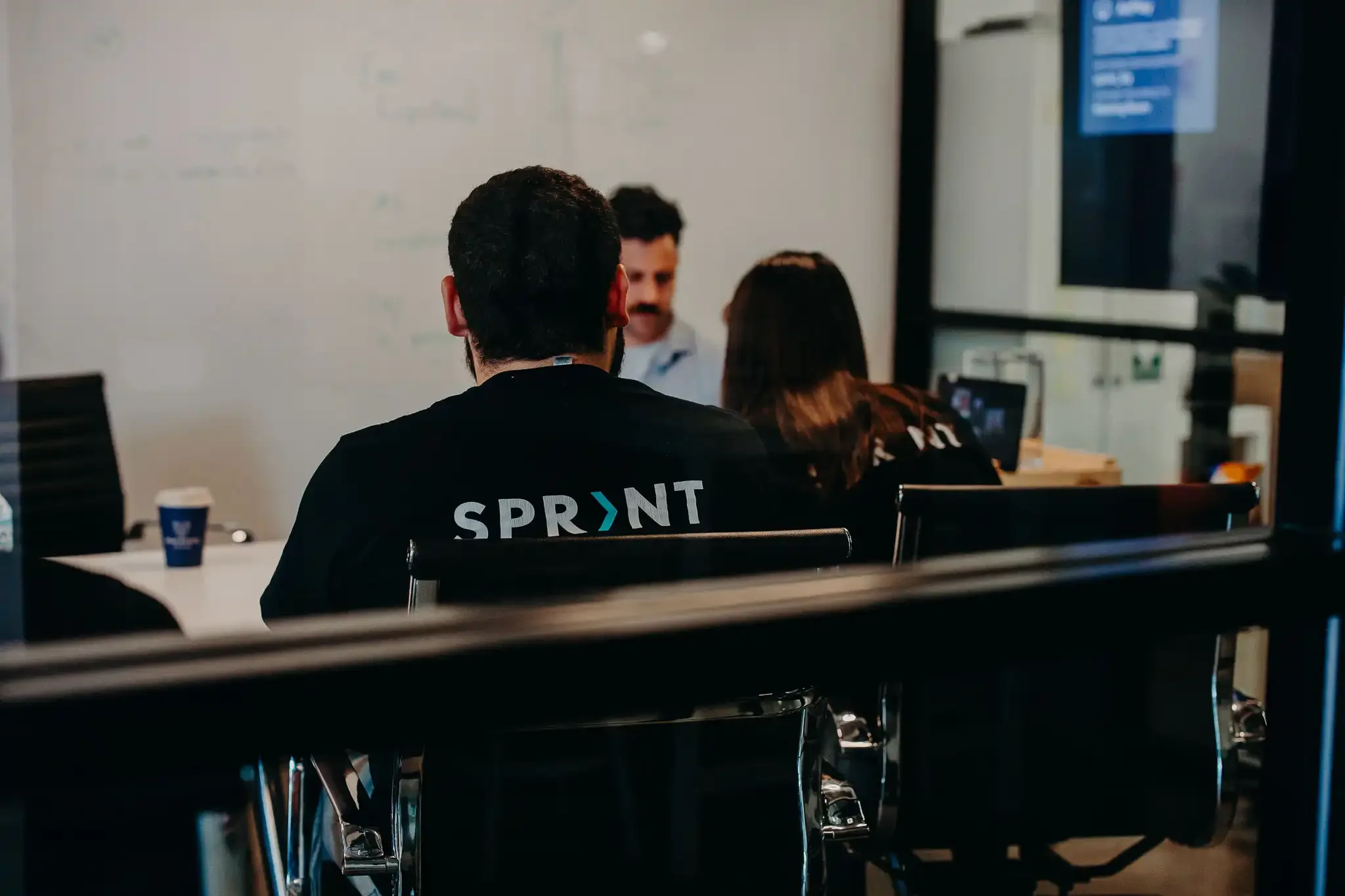 3 people in a meeting room, the photo is taken through the glass walls. 1 person is wearing a shirt with the word Sprint on the back of it, and the other 2 are engaged in a conversation. 