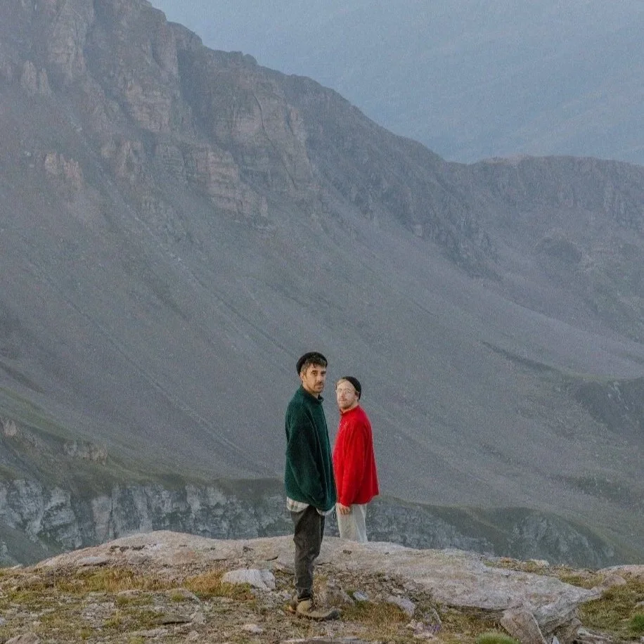 Two men wearing colorful jackets stand on a rock in a mountainous landscape.