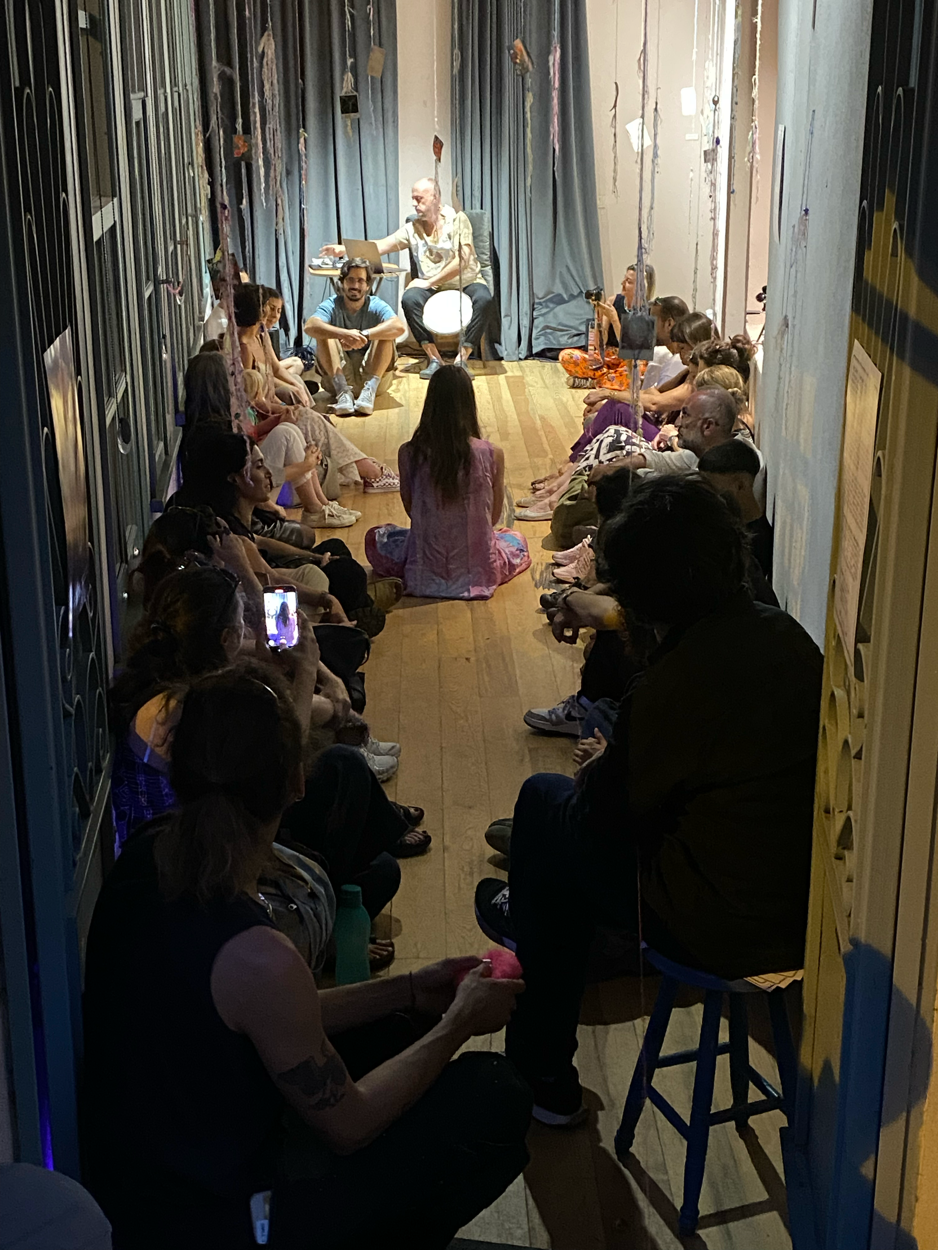 Une salle de spectacle avec une audience assise au sol, regardant une scène où deux personnes sont en train de performer, entourées de décorations suspendues.