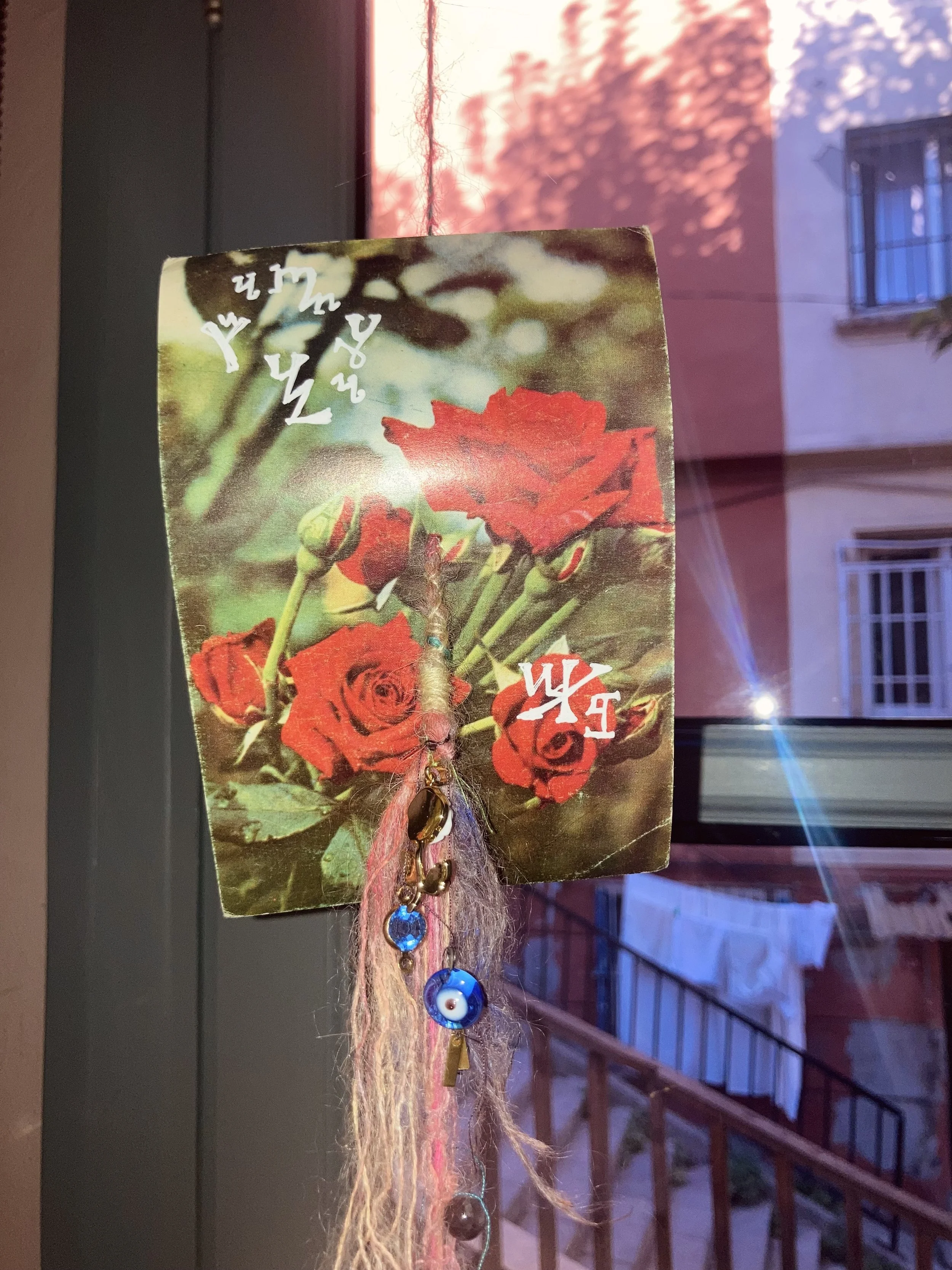 Une branche avec un collier de perles et charmes, accroché à une carte avec des roses rouges, en face d'une fenêtre avec vue sur un bâtiment