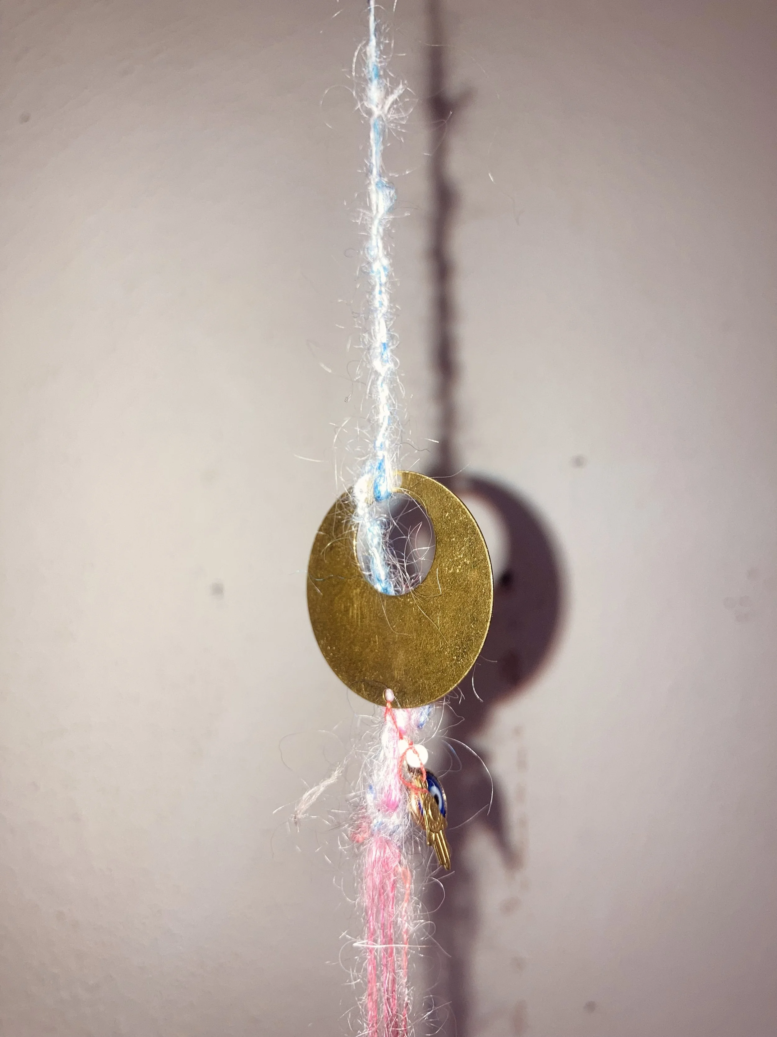 Une pièce de monnaie dorée suspendue à un fil de coton blanc, avec un petit porte-clés et un fil rose dessous, contre un mur clair.