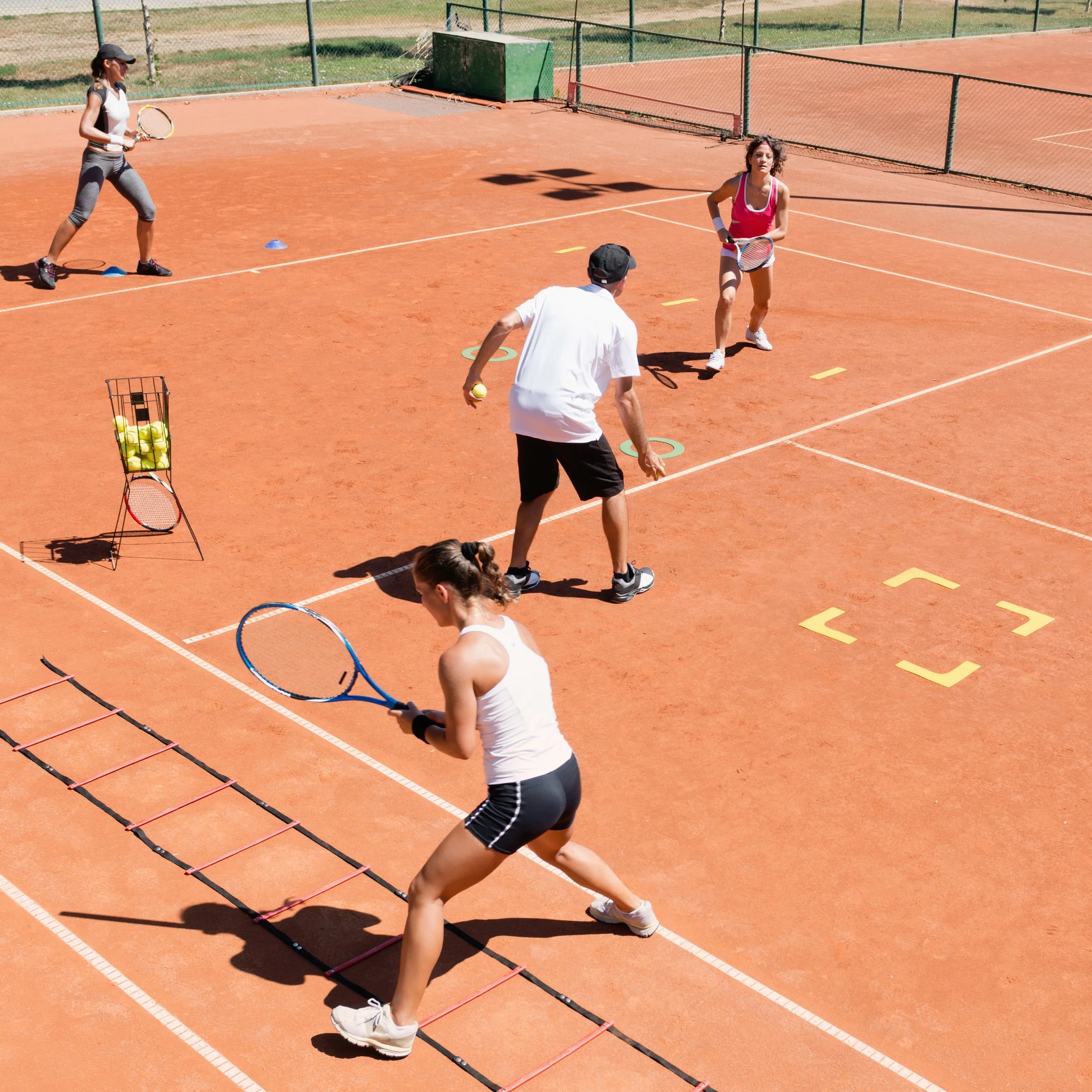 Vier Personen beim Tennis-Trainings auf einem roten Sandplatz, drei Frauen und ein Mann, alle mit Tennisschlägern, zwei Frauen bereiten sich auf den Schlag vor, eine Frau führt ein Koordinationstraining mit einem Leiter durch, im Hintergrund Tennisnetz und Trainingsgeräte.