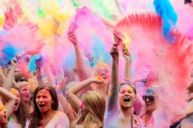 People celebrating with colored powder at a Holi festival, throwing vibrant pink, yellow, green, blue, and red powders into the air.