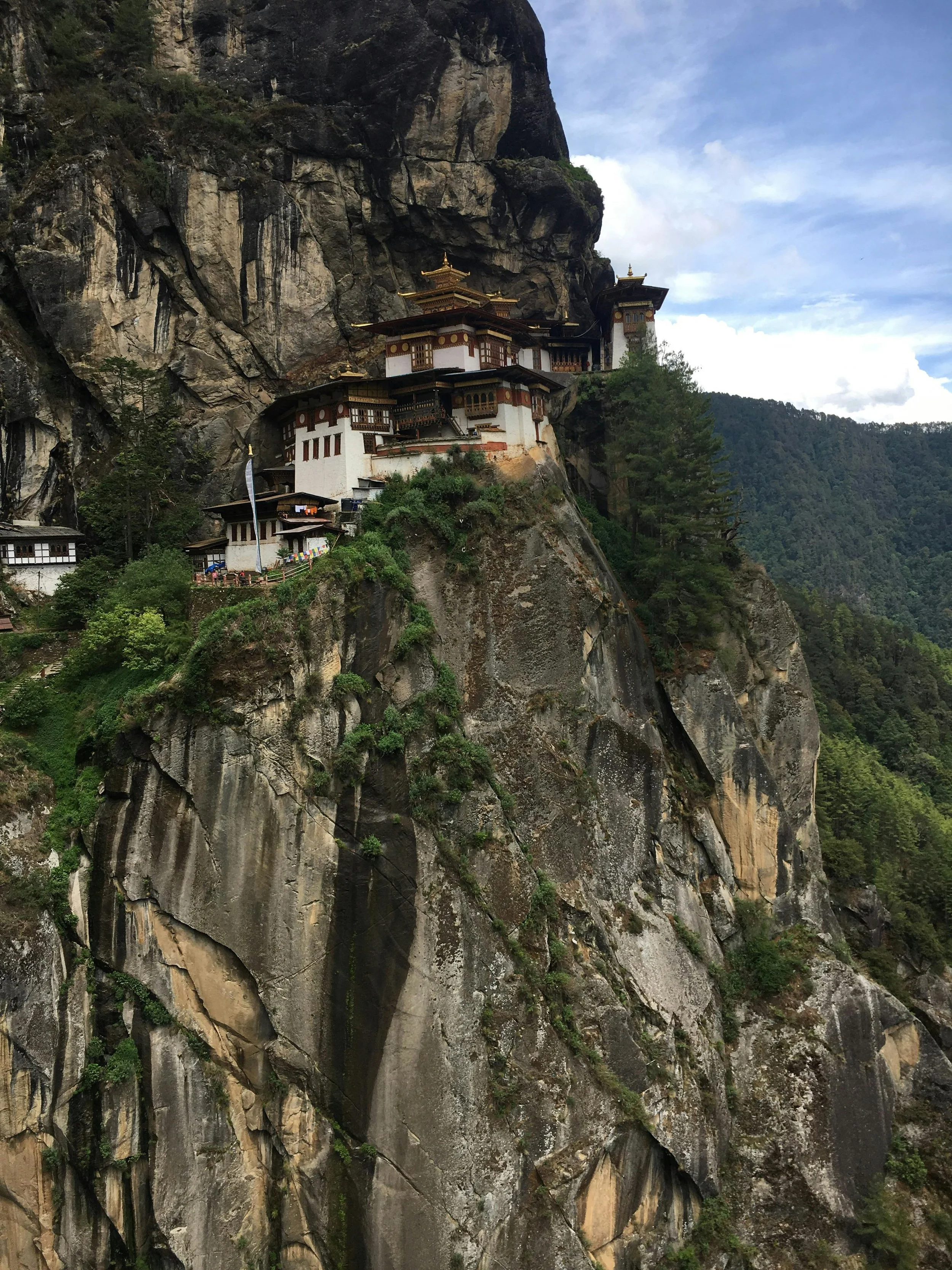 A mountain monastery built on a steep cliff, surrounded by trees and overlooking a forest, under a partly cloudy sky.