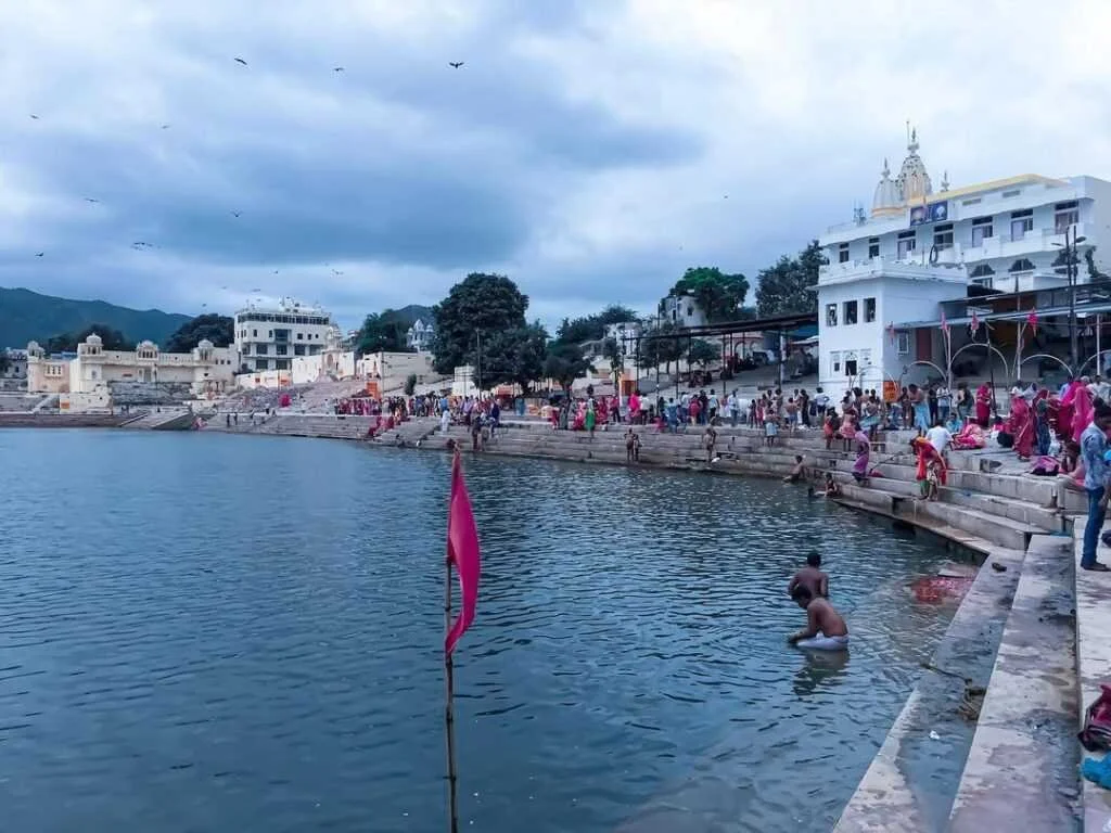 A busy ghats along a river, with people bathing and gathering, white buildings, and hills in the background, under cloudy skies.