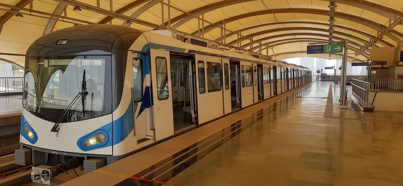 A modern train at an elevated station platform with an arched roof and city skyline in the background.