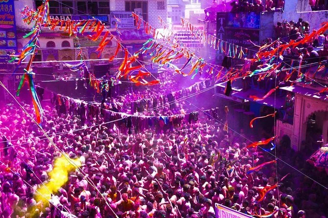 A large crowd celebrating in the streets with colorful decorations and streamers, yellow smoke, and purple lighting.