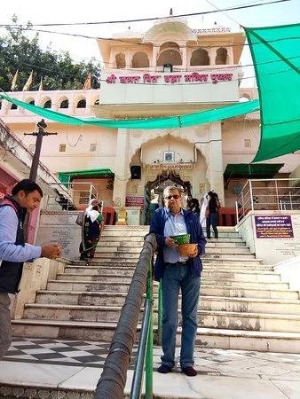 People standing outside a white temple with pink decorations and a dome, stairs leading up to the entrance, green canopy, and a large sign with Hindi text