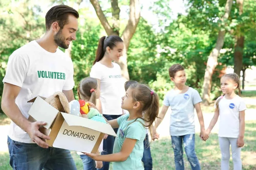 A man and woman in volunteer T-shirts distributing donations to children outdoors, with trees and grass in the background.