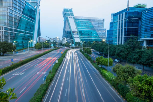 Nighttime cityscape with modern glass buildings and light trails from moving vehicles on a multi-lane road in an urban area.