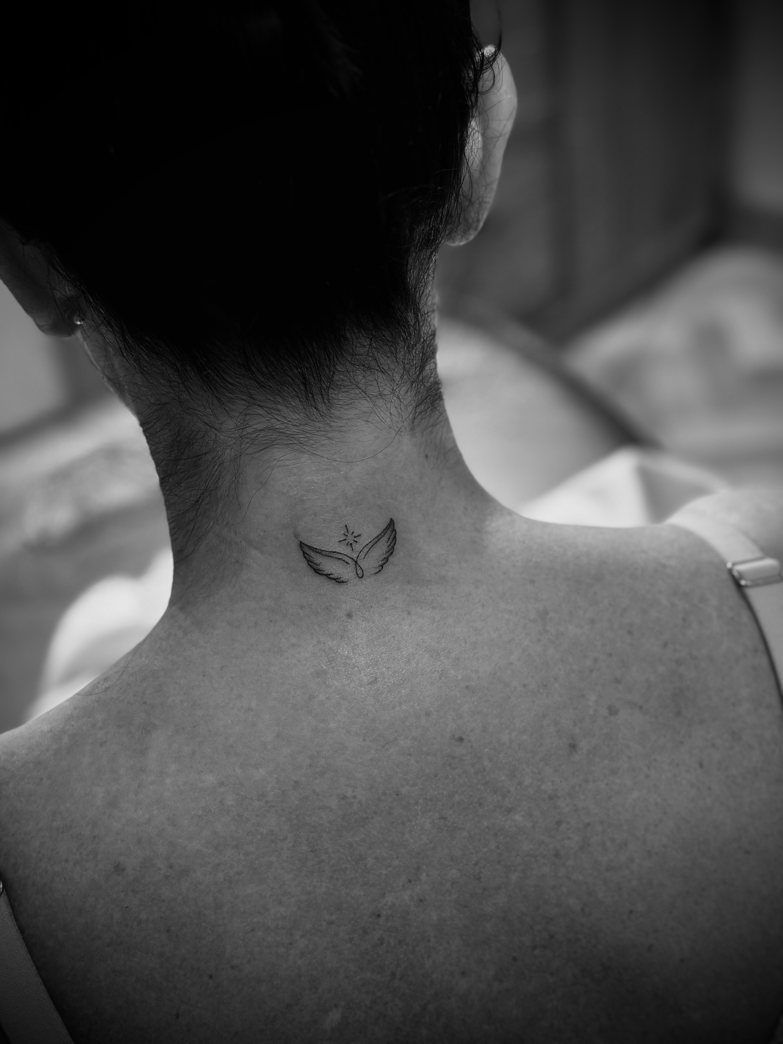Black and white photo of a small tattoo on the back of a person's neck, featuring a design with curved wings and a star above it.