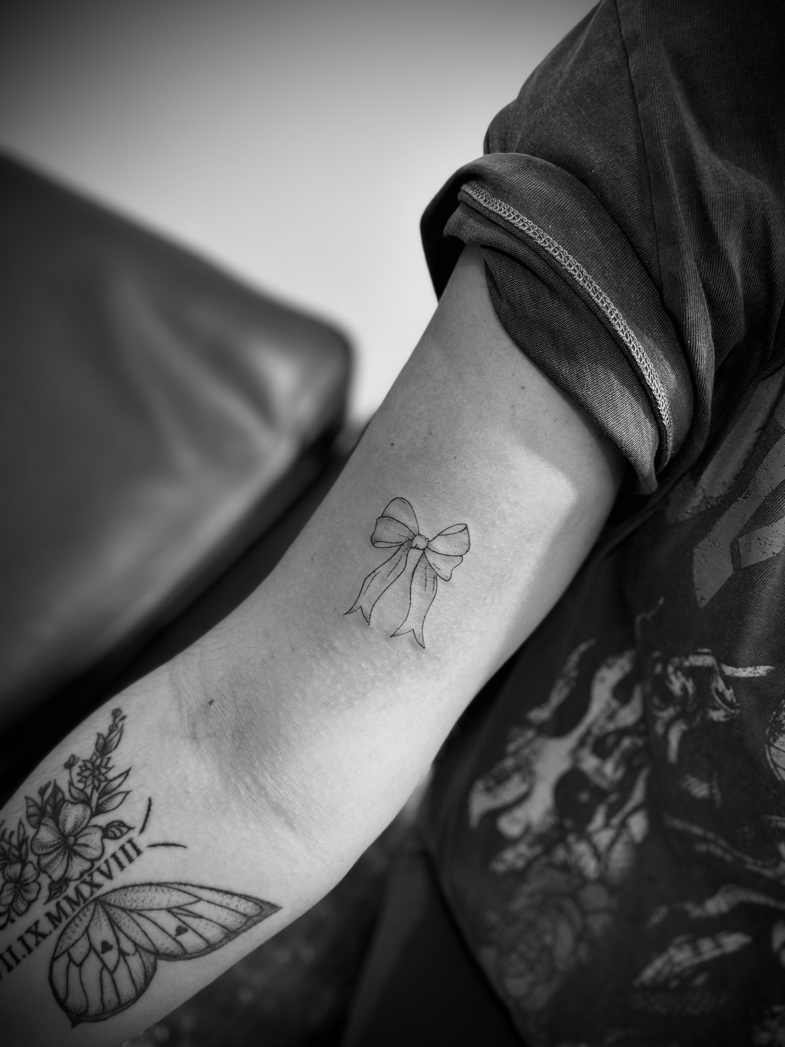 Arm with multiple tattoos, including a bow design and floral pattern with Roman numerals, in black and white.