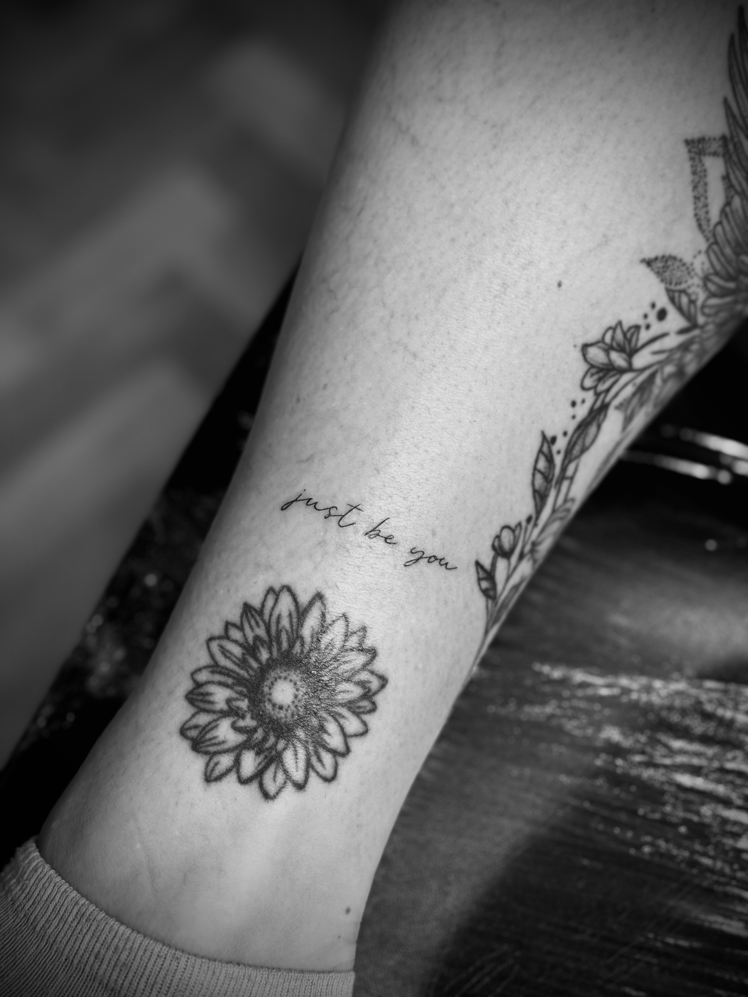 Black and white tattoo on a person's leg featuring a sunflower and the text "just be you" accompanied by floral accents.