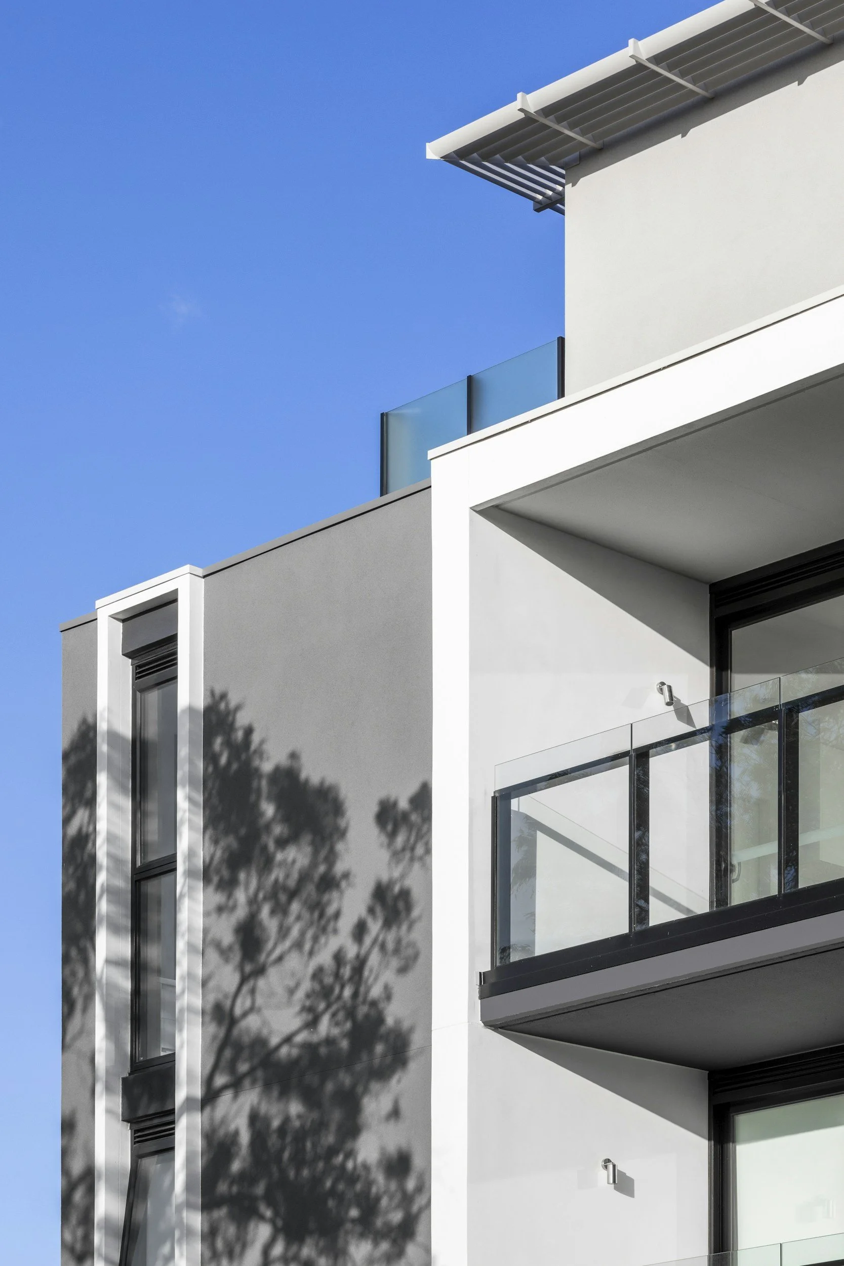 Moderne weiße und graue Wohnblock mit Balkonen, Schatten eines Baumes an der Fassade und blauer Himmel.