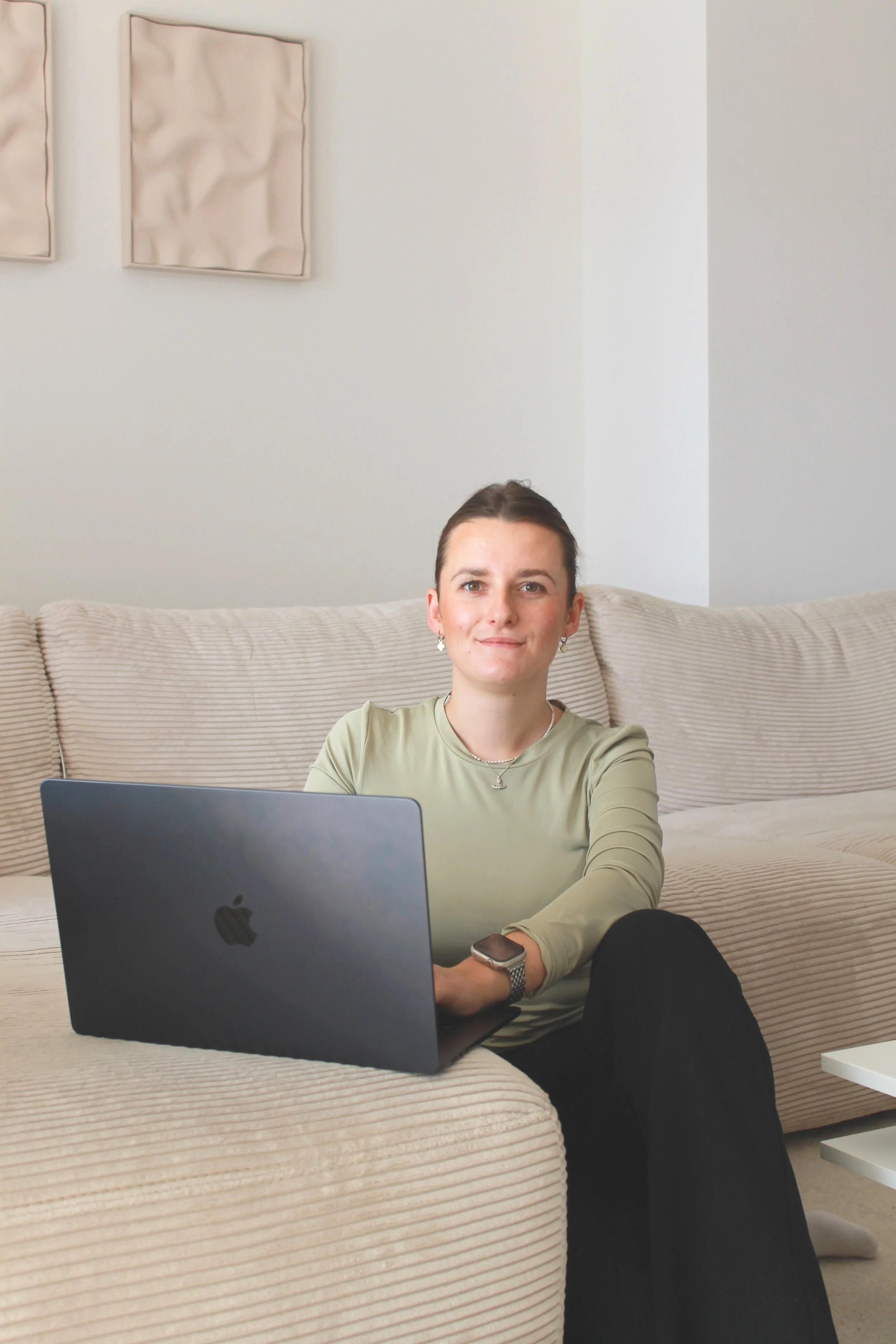 Junge Frau sitzt auf einer Couch vor einem Laptop, trägt eine Uhr und sieht die Kamera an, in einem modernen, hellen Wohnzimmer.