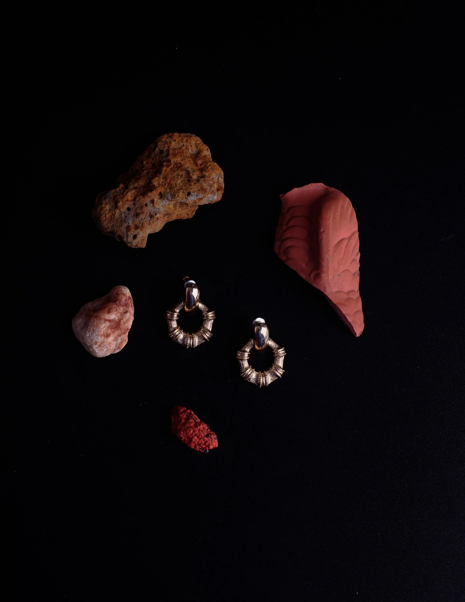 Various rocks and jewelry pieces displayed on a black background, including a textured rust-colored rock, a pinkish-red geode, smaller stones, and metallic hoop earrings.