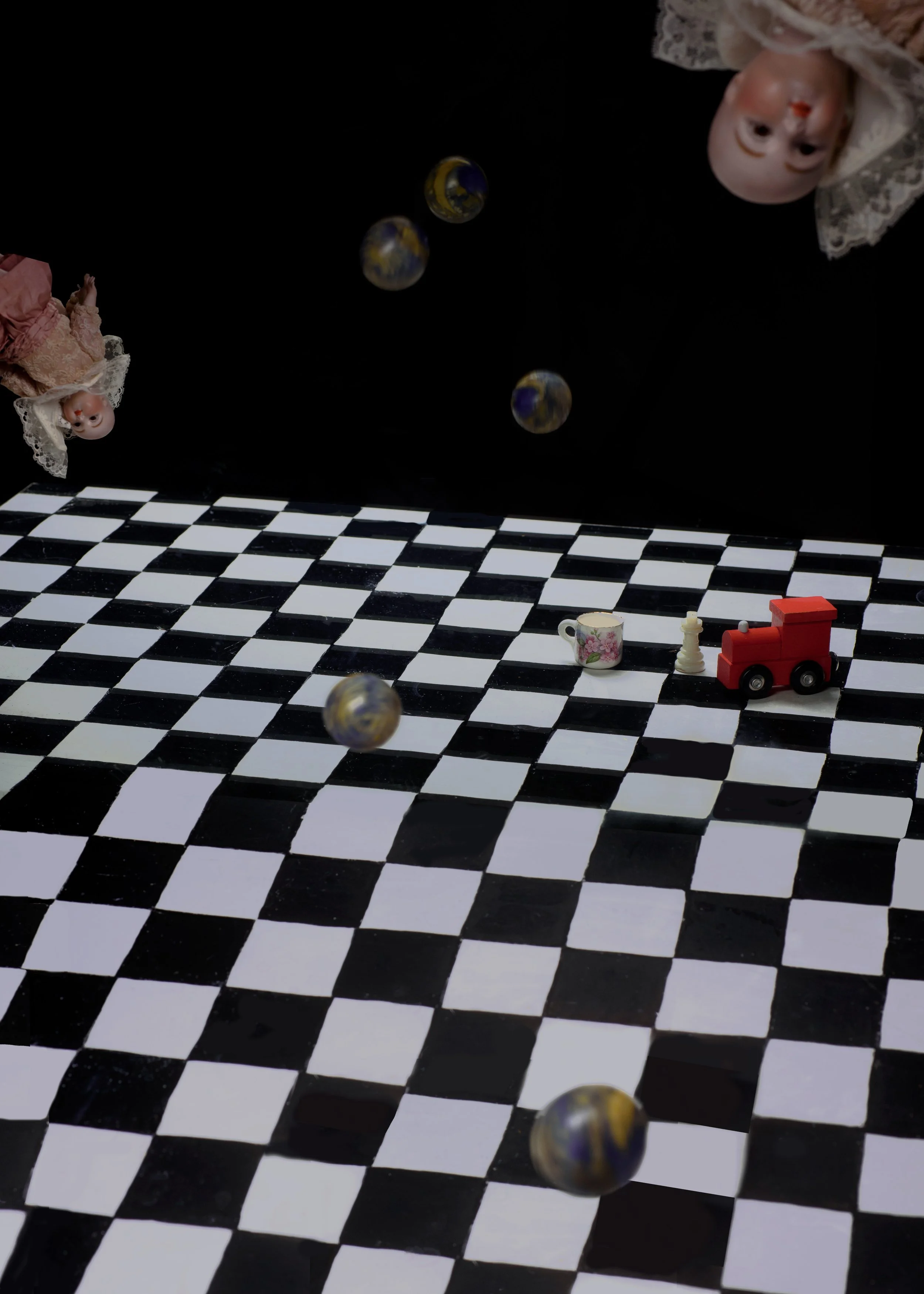 Dolls' heads with lace head coverings, a checkered black and white floor, and small globe-like objects floating in front of a black background. There is also a toy red train, a floral teacup, and a white chess piece on the checkered floor.