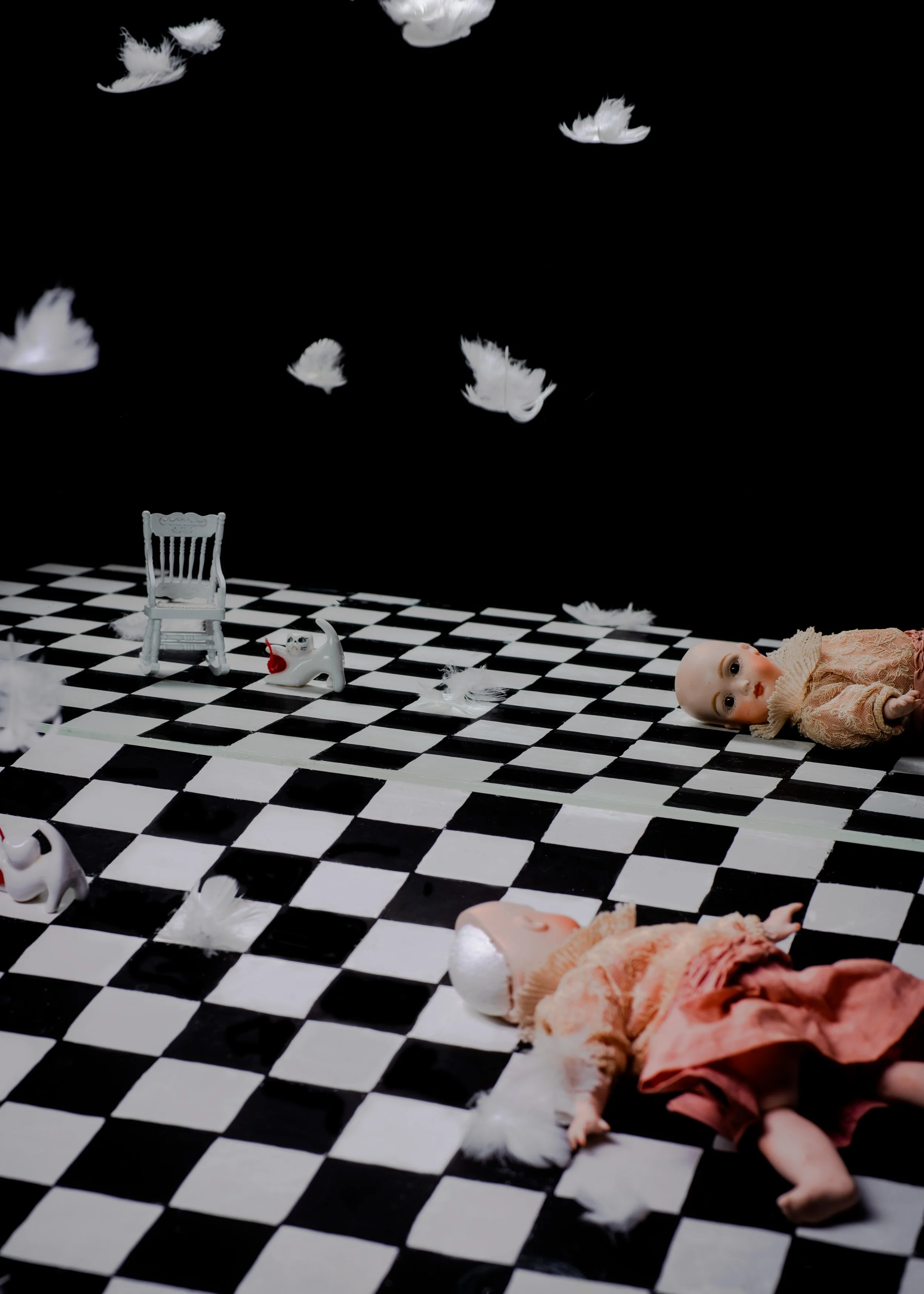 A doll lying on a black and white checkered floor, with scattered white feathers, small white ceramic figures of cats, a white chair, and a partly visible doll in a pink dress.