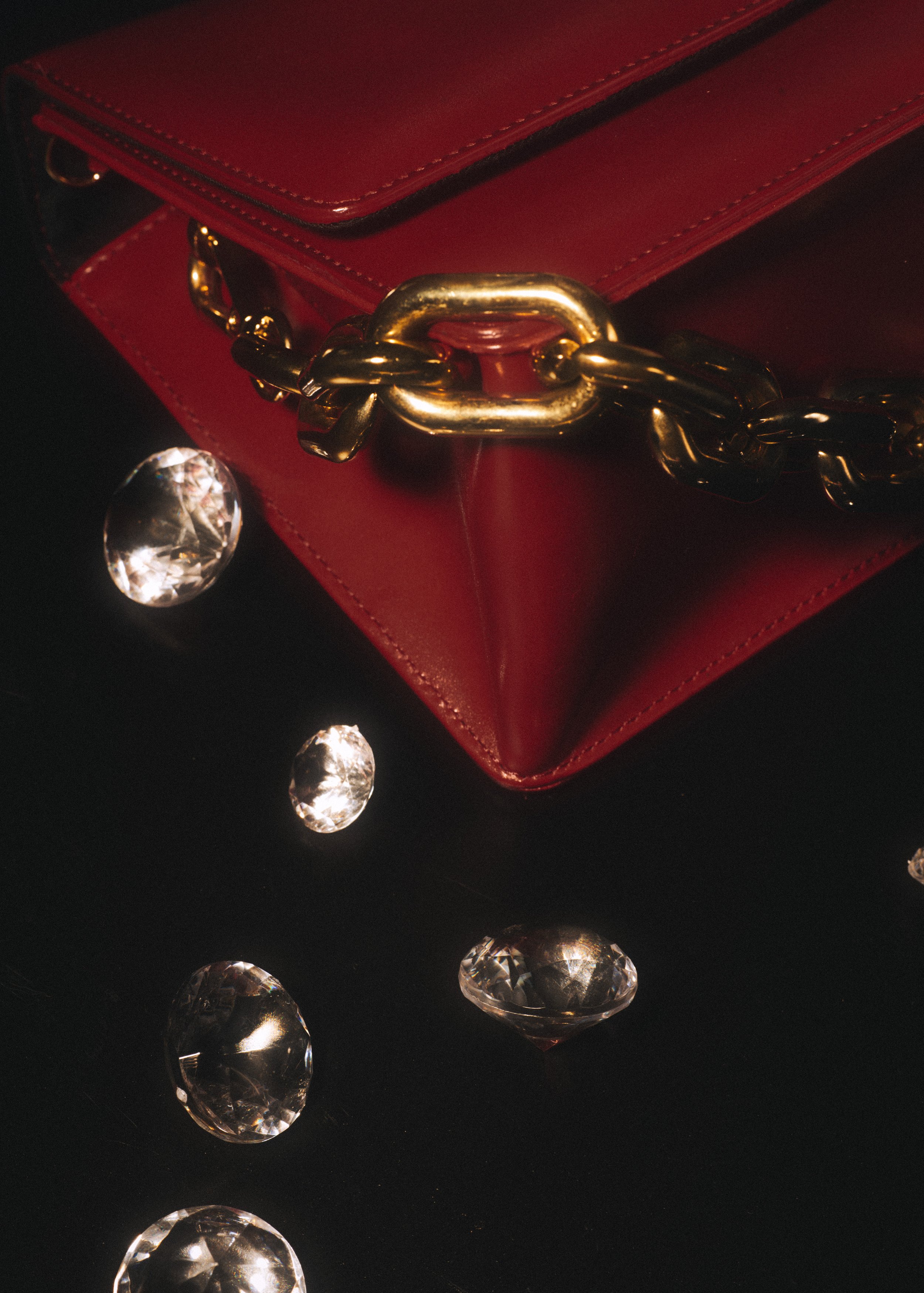 A red handbag with a gold chain strap and a gold clasp, surrounded by scattered clear gemstones on a black surface.