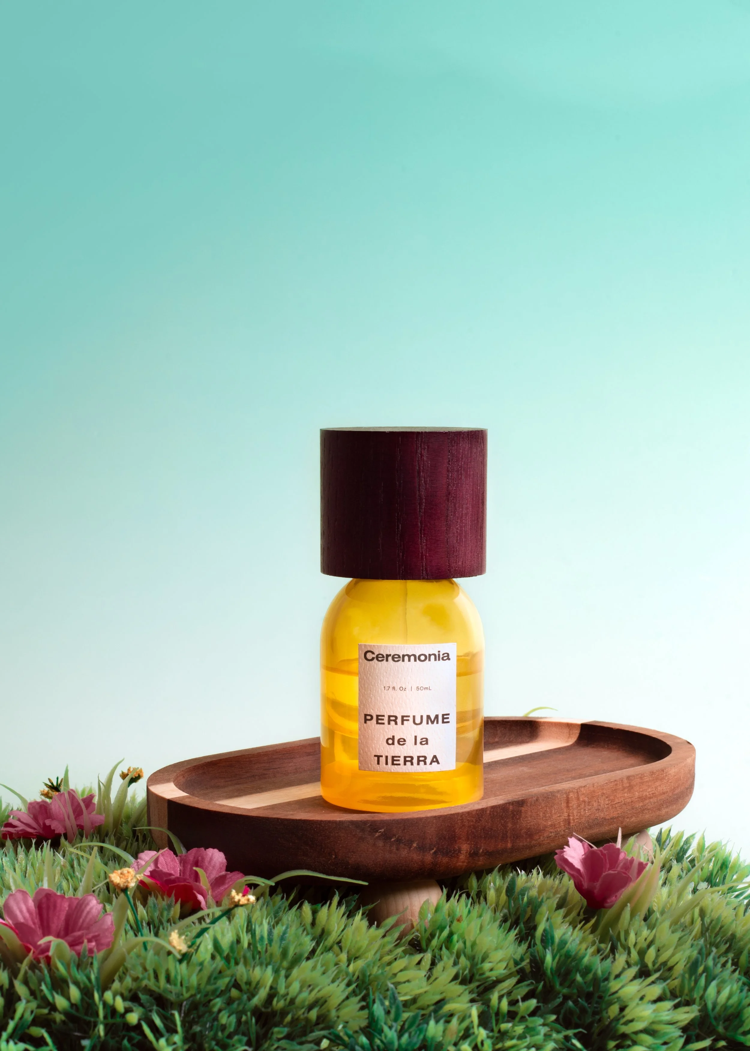 A yellow perfume bottle labeled "Ceremonia PERFUME de la TIERRA" with a dark red cap, placed on a wooden tray surrounded by pink flowers and green grass against a light blue background.