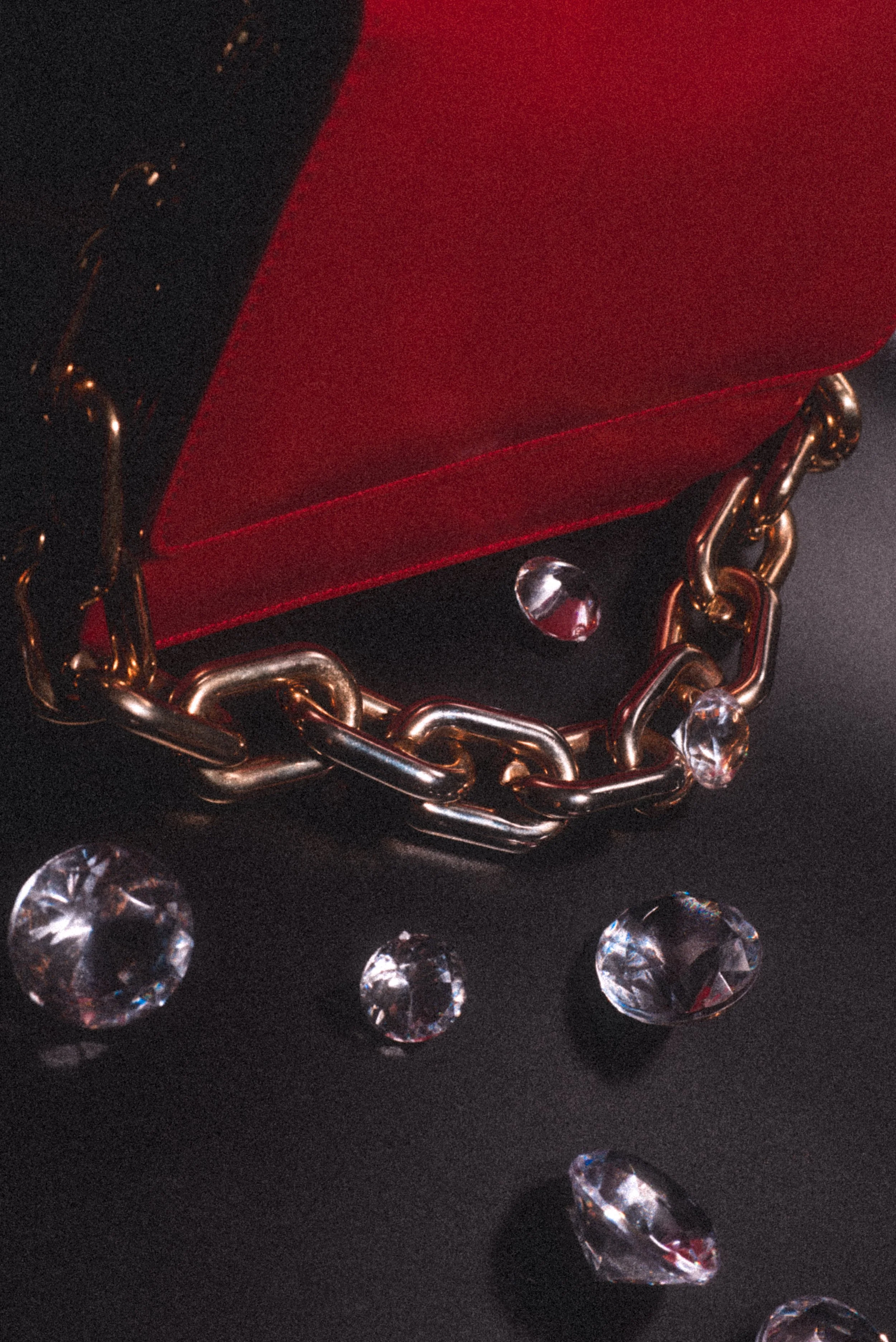 A red jewelry box with a gold chain chain and scattered clear, shiny gemstones on a black surface.