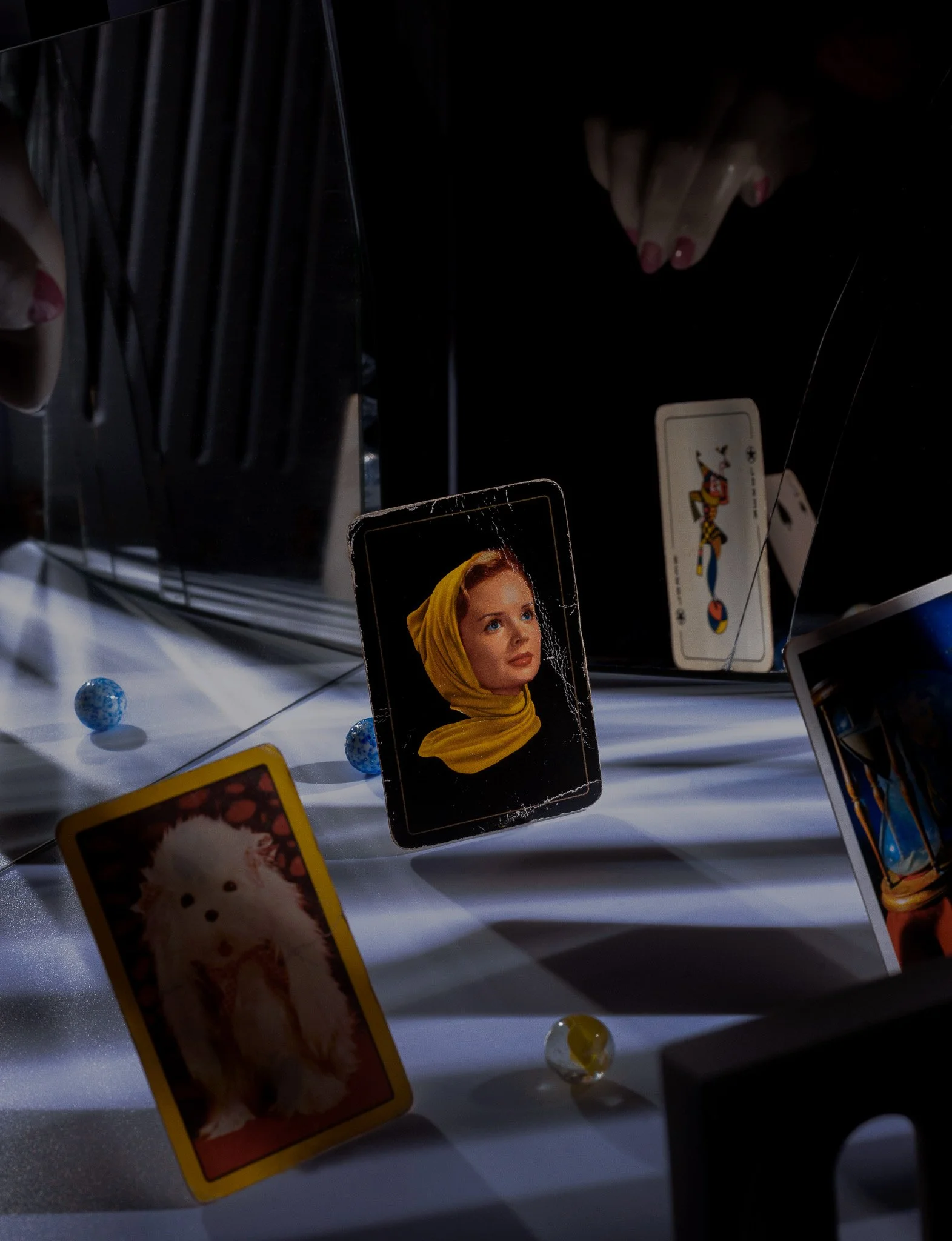 A reflection in a mirror shows playing cards, including a card with a portrait of a woman in a yellow headscarf, a card of jester, and a card of a dog, on a surface with small decorative balls.
