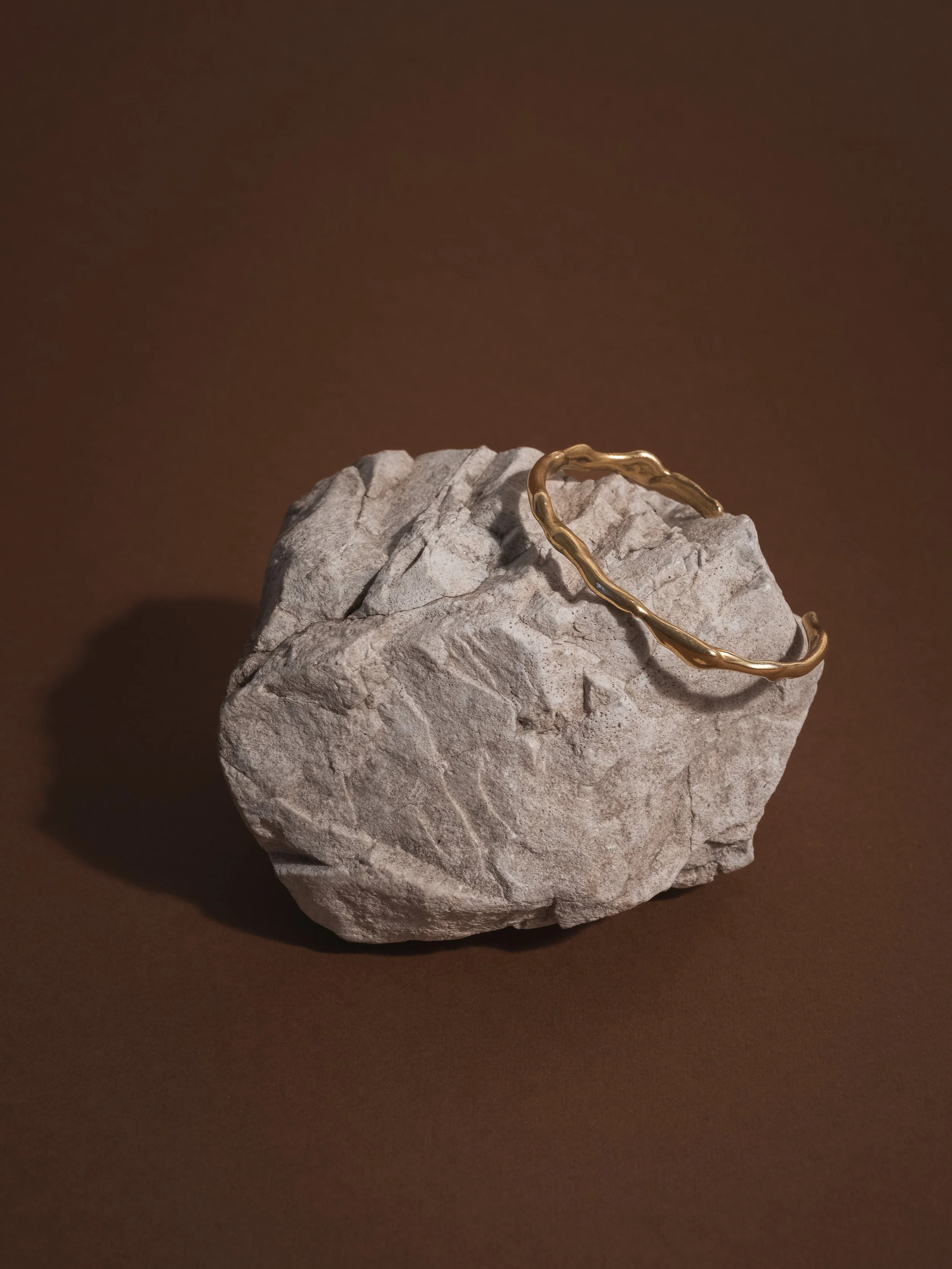 Bracciale rigido di colore oro, posizionato su una roccia di pietra grigia, sfondo marrone.