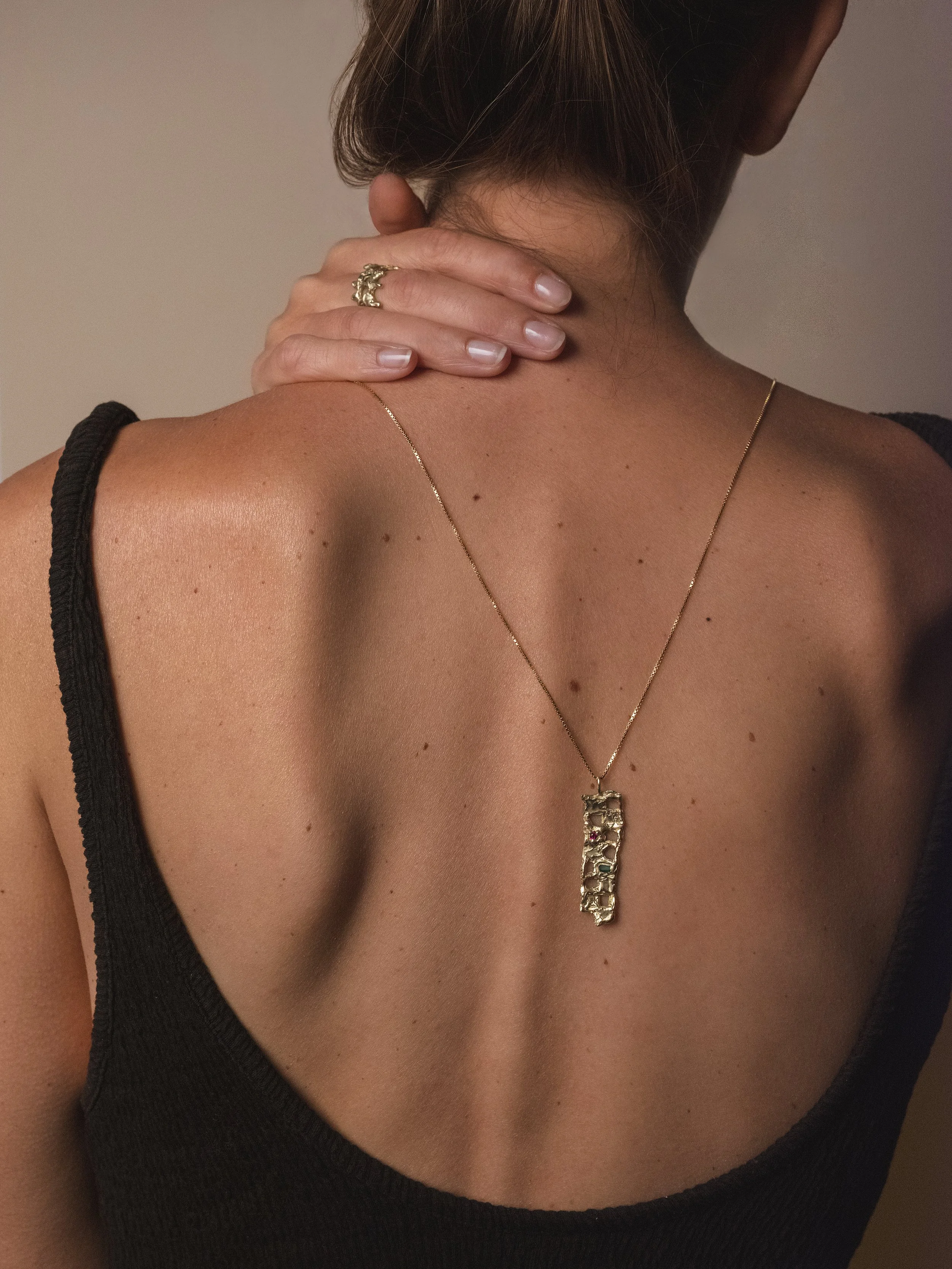 Parte superiore del corpo di una donna con capelli lisci che indossa una collana dorata con pendente e un anello sulla mano destra, che si sistema sulla pelle del collo e della spalla.