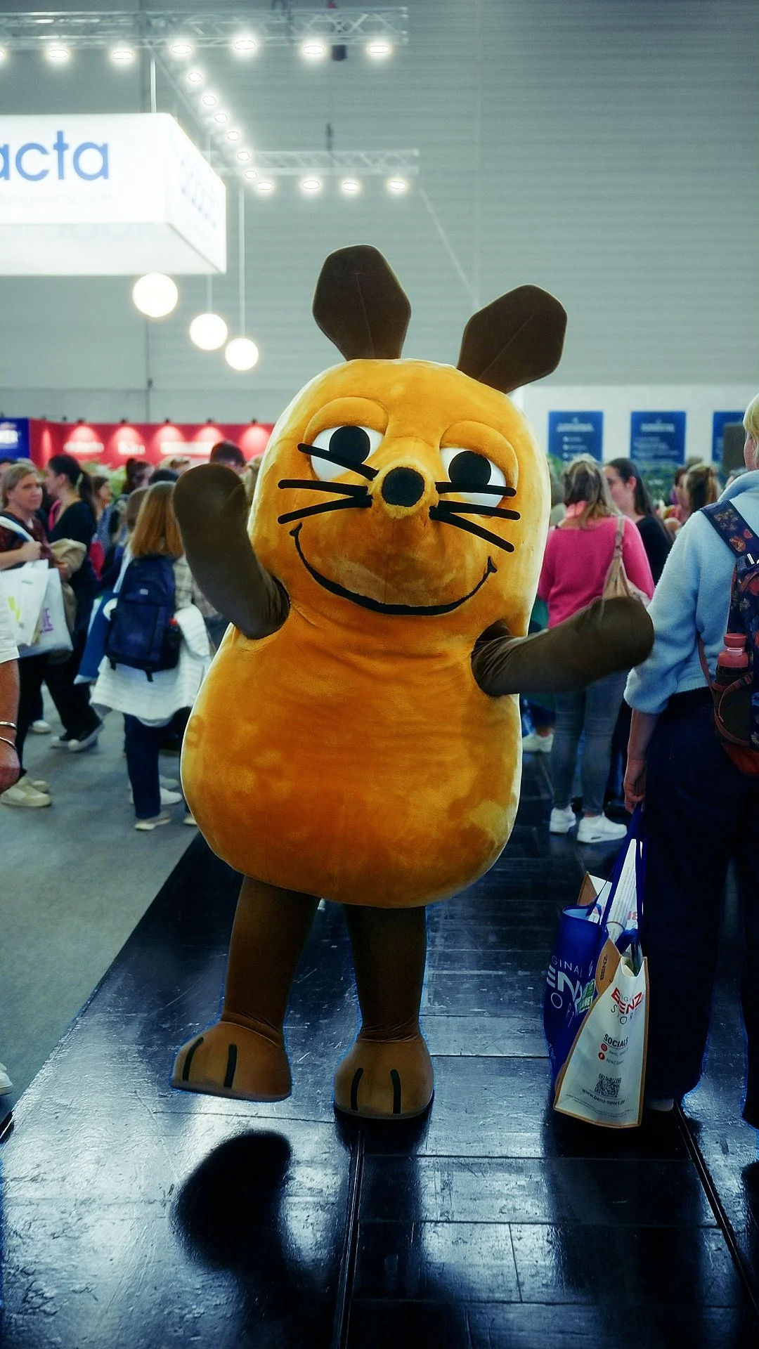 Costumed character in a bunny costume with a orange body and brown ears, smiling and waving at a crowded indoor event.