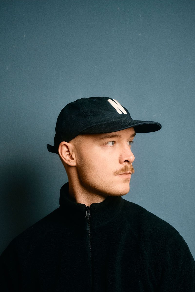 Junge Mann trägt schwarze Mütze und schwarzen Pullover, blickt nach rechts, vor einer blauen Wand.