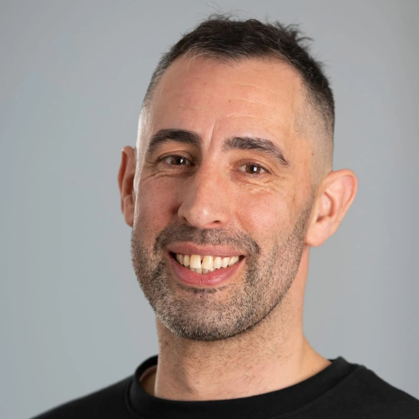 Smiling man with short hair and beard, wearing a black shirt, against a gray background.