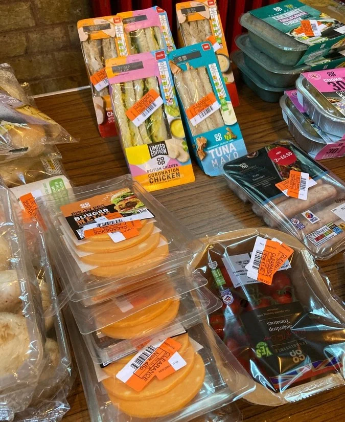 Assorted pre-packaged sandwiches, chicken Caesar wraps, tuna mayo sandwiches, vegetable salads, and sliced fruit in plastic containers with orange price tags on a wooden table.