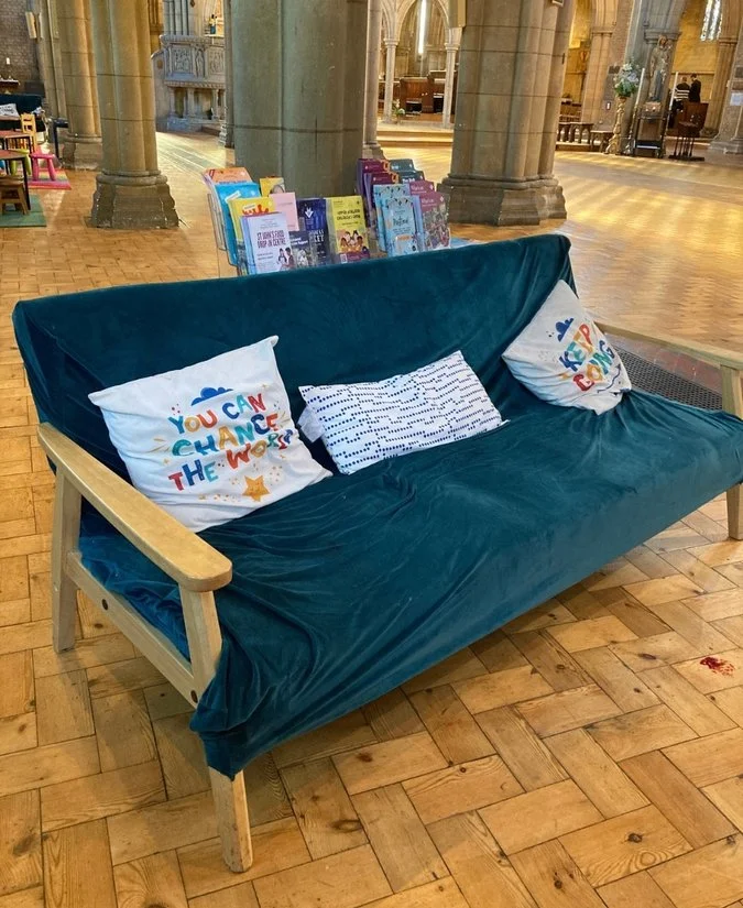 A blue velvet sofa with wooden armrests, three decorative pillows, and a bookshelf filled with children's books in a church or cathedral interior.