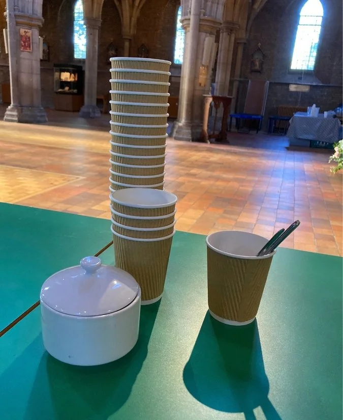 A stack of disposable cups, a single disposable cup with utensils, and a small ceramic container with a lid on a green table inside a church.