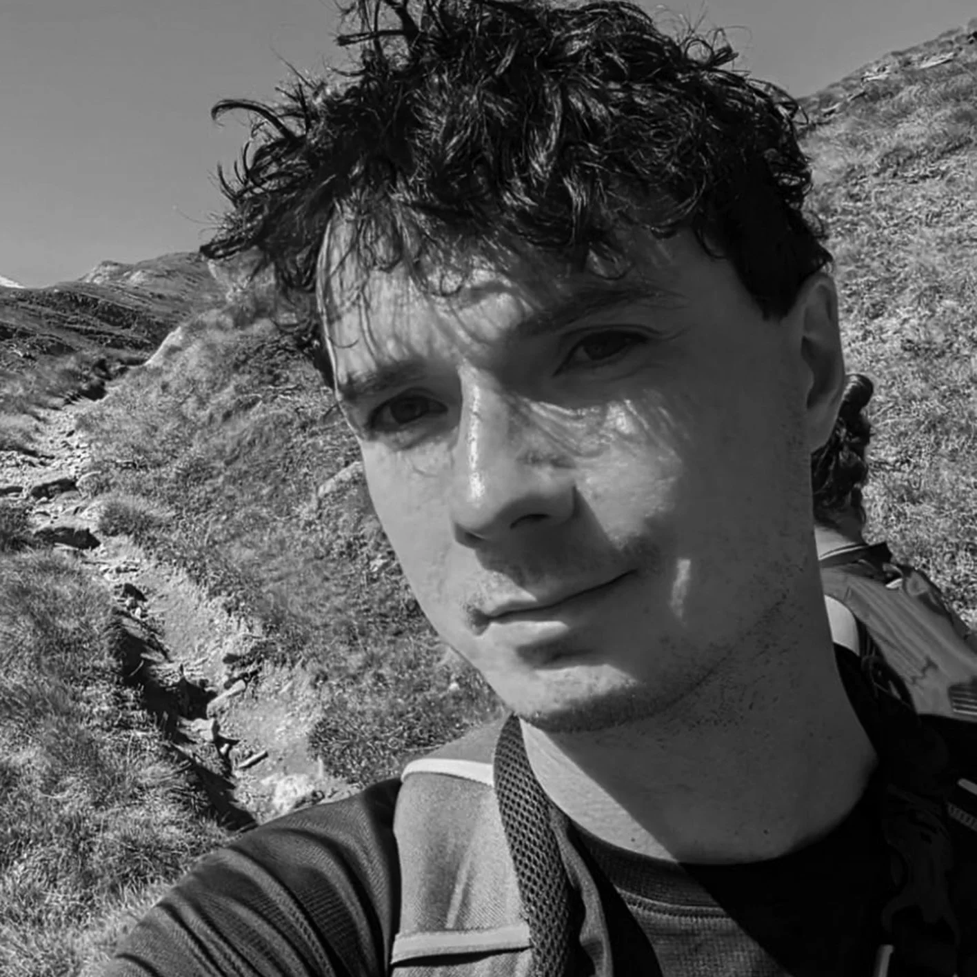 A black and white photo of a person outdoors, wearing a backpack. They are on a trail surrounded by grassy hills.