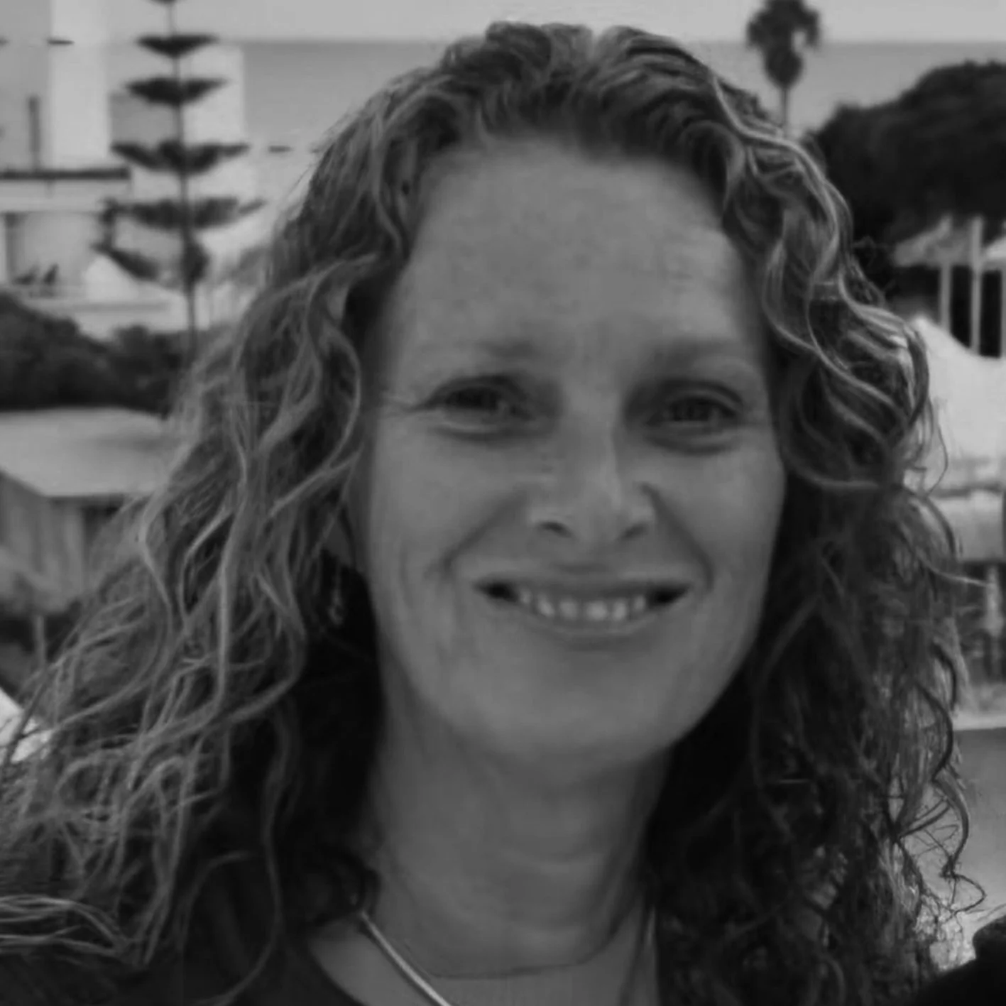 A grayscale photo of a woman with curly hair smiling outdoors, with trees and buildings in the background.