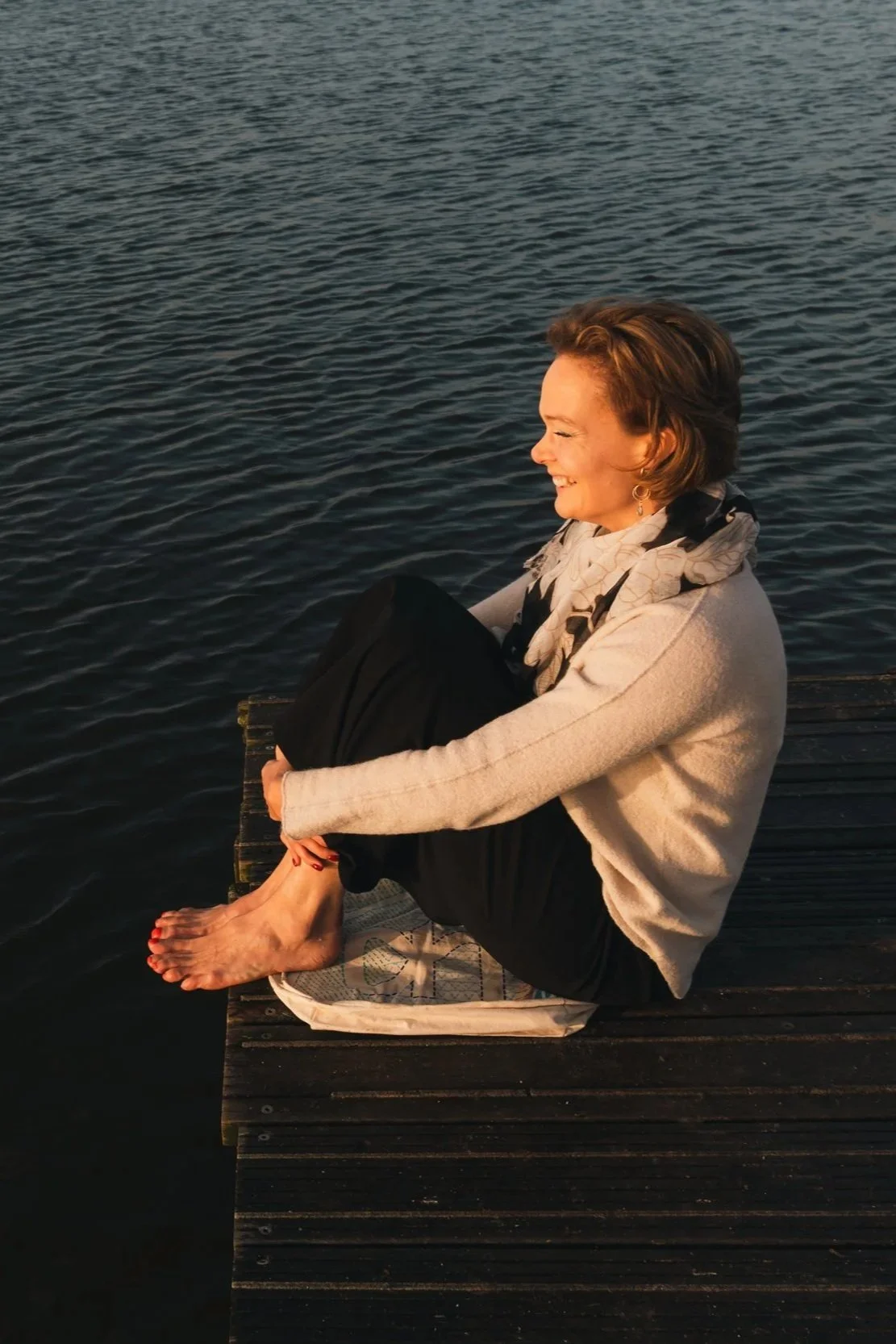 Vrouw zit op een houten steiger aan het water, glimlacht in de zon