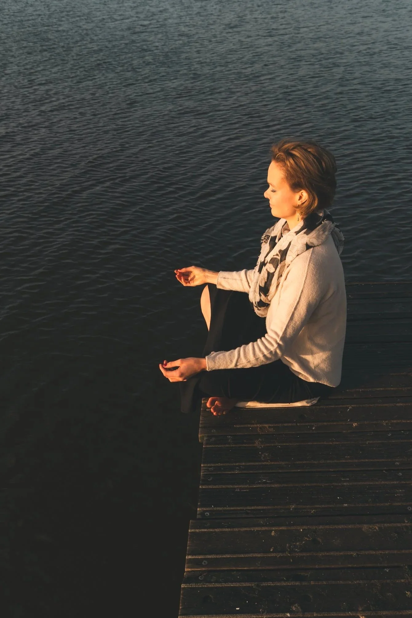 Vrouw zit in kleermakerzit op houten steiger naast een meer, kijkt ontspannen naar het water tijdens zonsondergang.